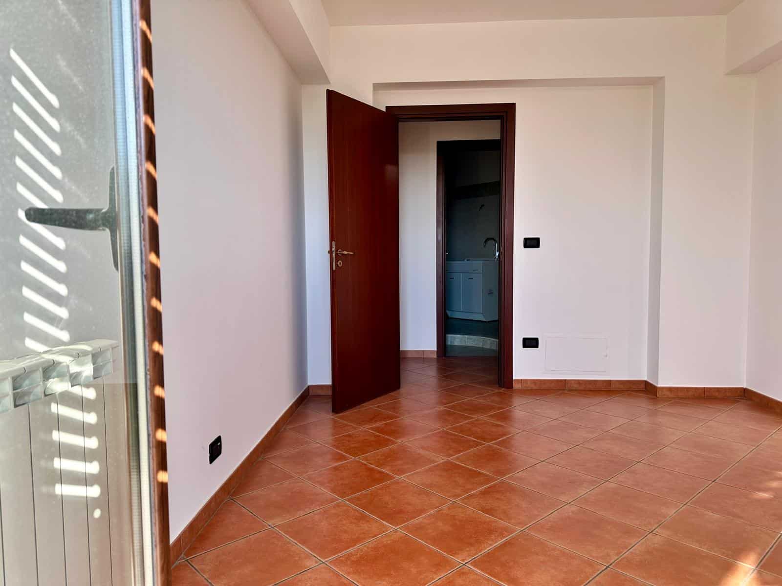Ampia stanza luminosa con pareti bianche e pavimento in piastrelle terracotta, porta aperta che conduce a un'altra stanza, luce naturale che entra dalla finestra a sinistra.