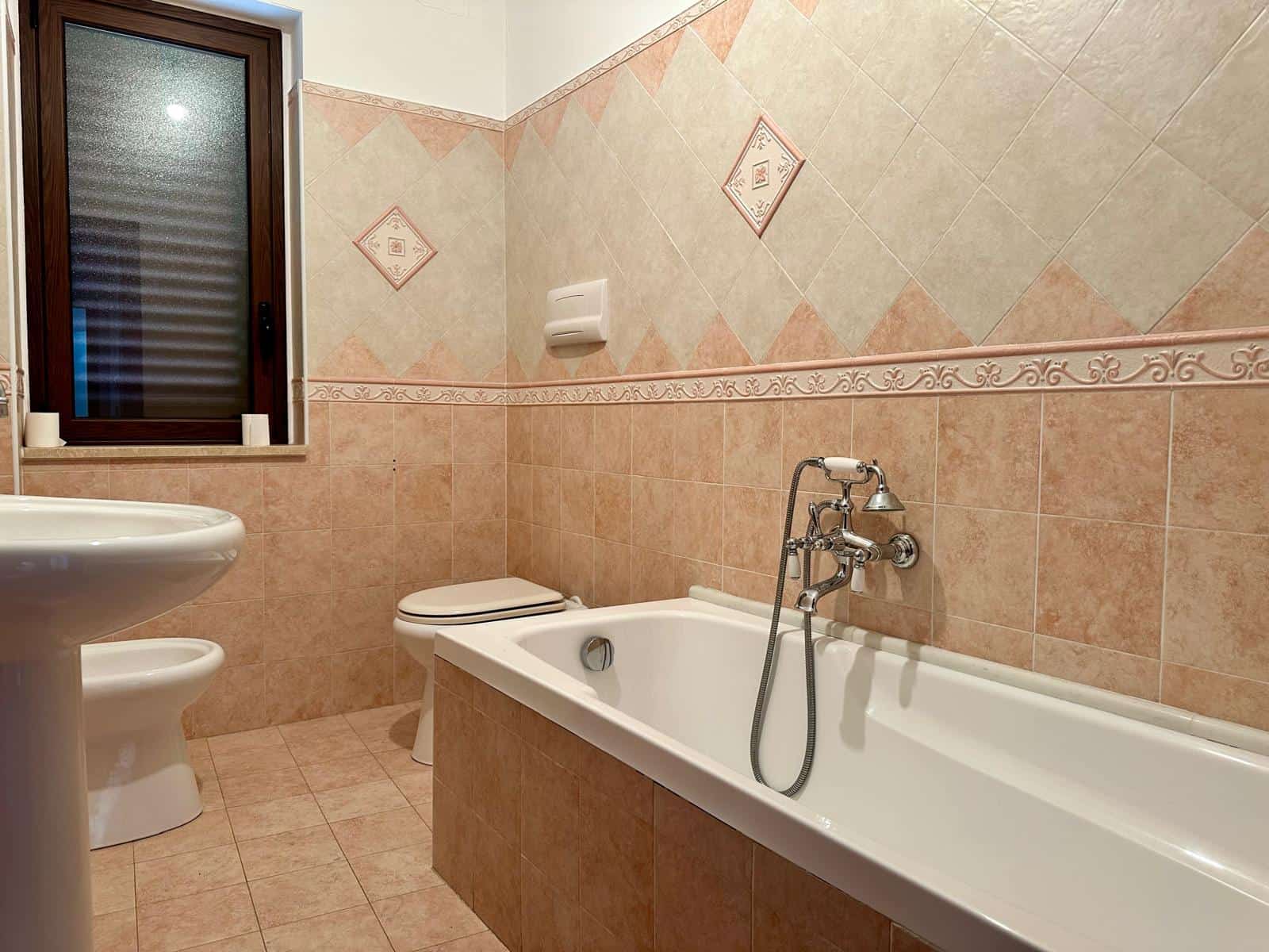 Bagno con vasca e finestra, rivestimenti in piastrelle beige e dettagli decorativi, ambiente luminoso e accogliente, ideale per il comfort domestico.