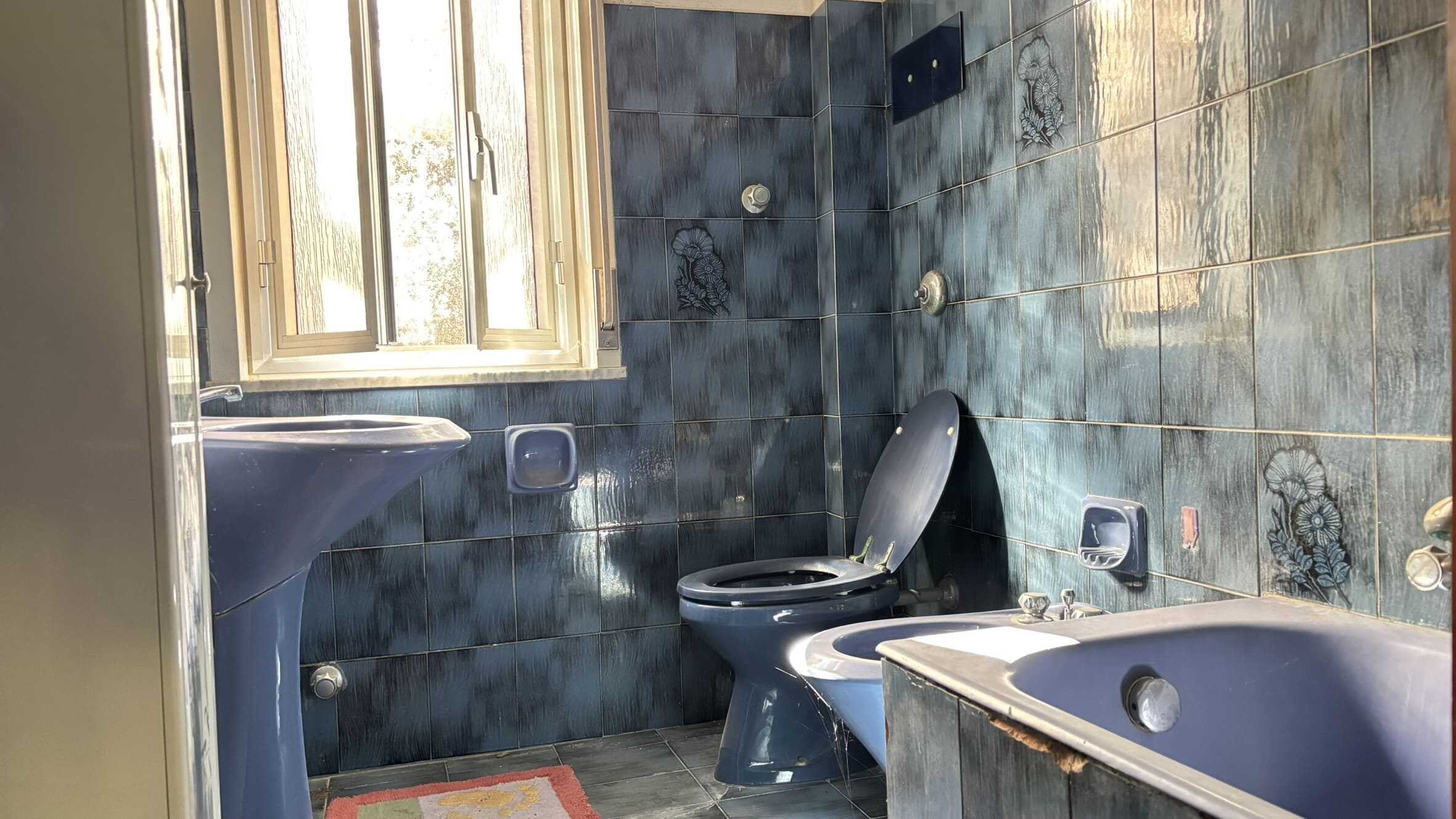 Lavabo, WC e vasca da bagno in bagno con pareti e pavimenti rivestiti in piastrelle blu con motivi floreali, finestra in legno che permette l'ingresso di luce naturale, ambiente classico e funzionale.