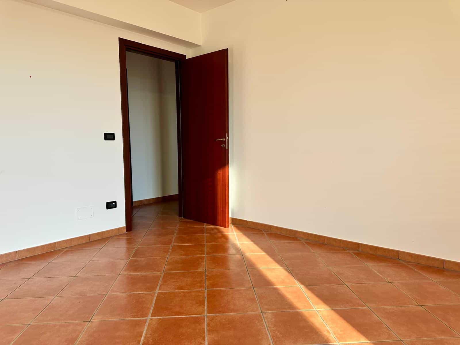 Stanza vuota con pareti bianche e porta in legno aperta, pavimento in piastrelle di terracotta, luce naturale che entra dalla finestra.