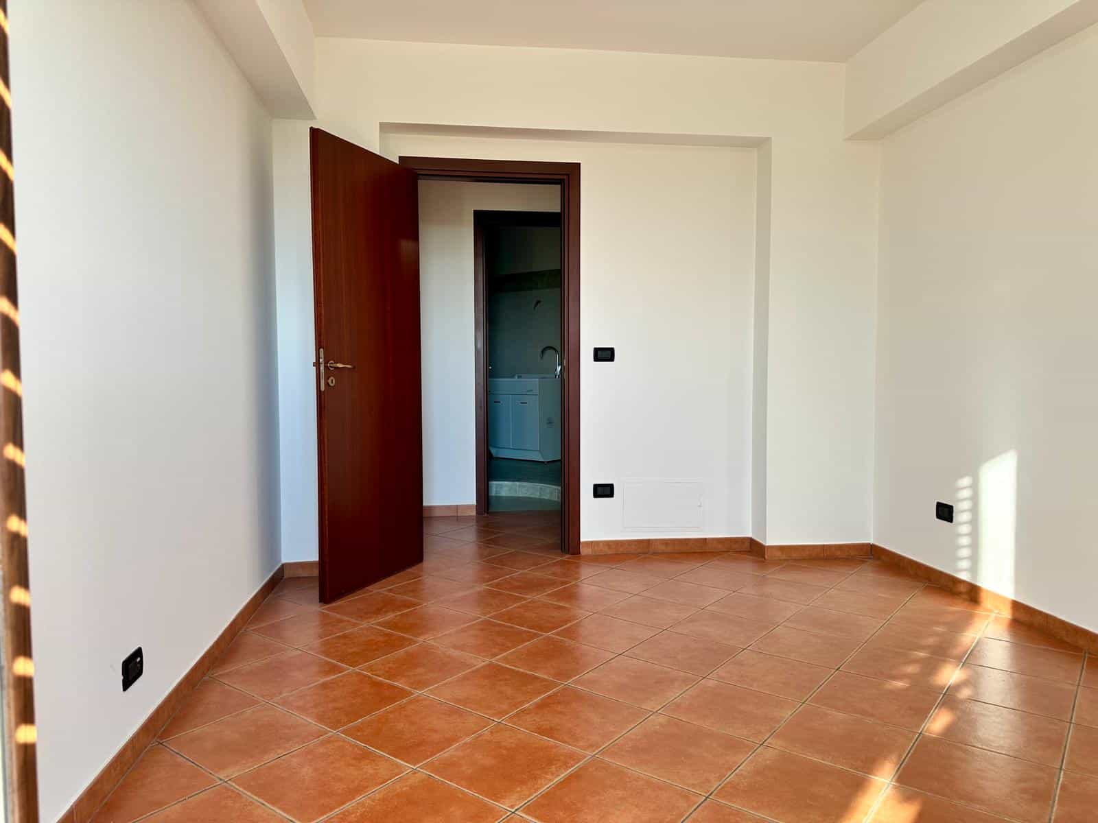 Stanza vuota con pareti bianche, pavimento in piastrelle arancioni e porta aperta che conduce a un'altra stanza, illuminata dalla luce naturale.