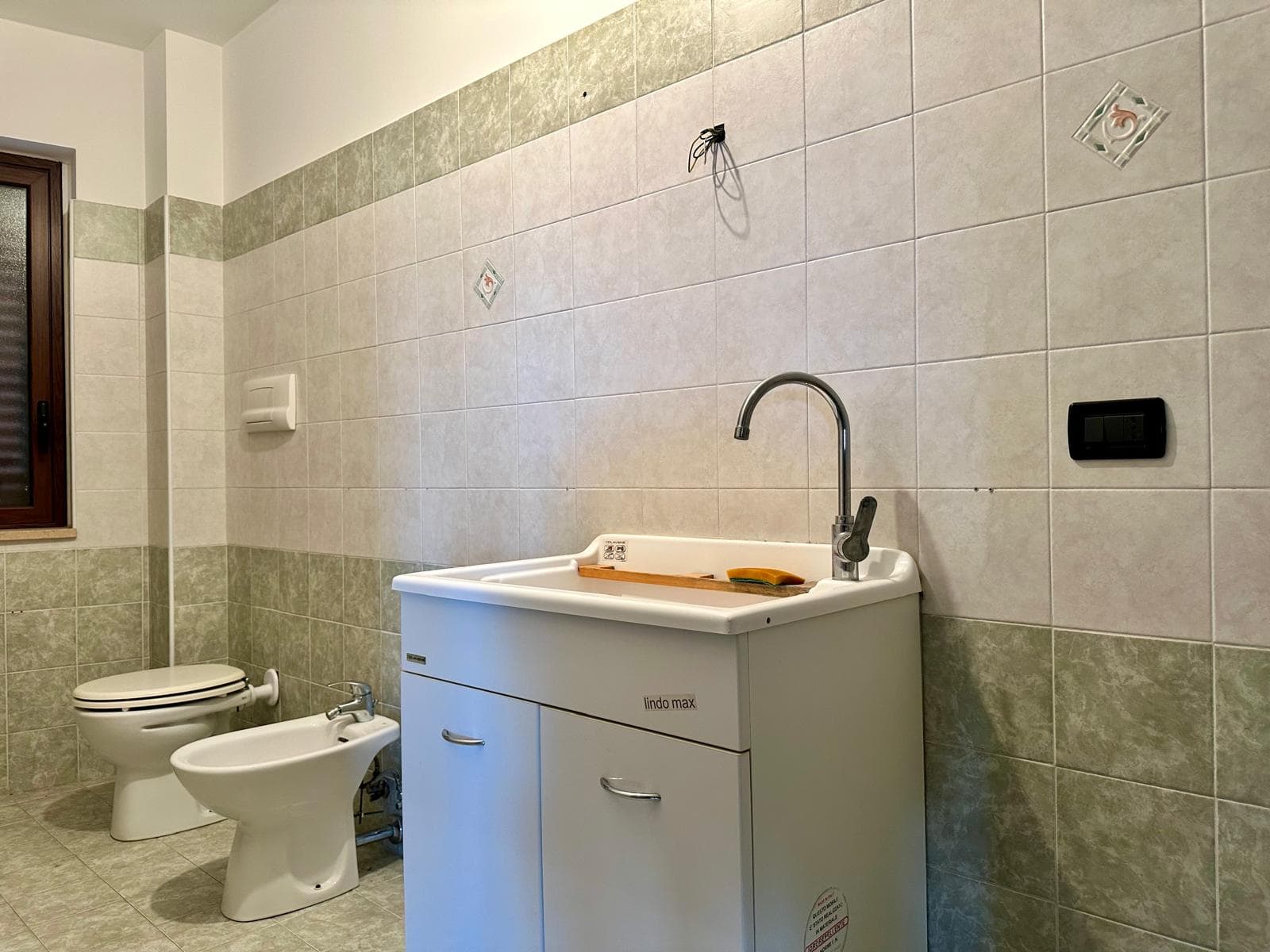 Lavabo e servizi igienici in bagno con piastrelle beige e verde, senza accessori visibili, ambiente pulito e funzionale.