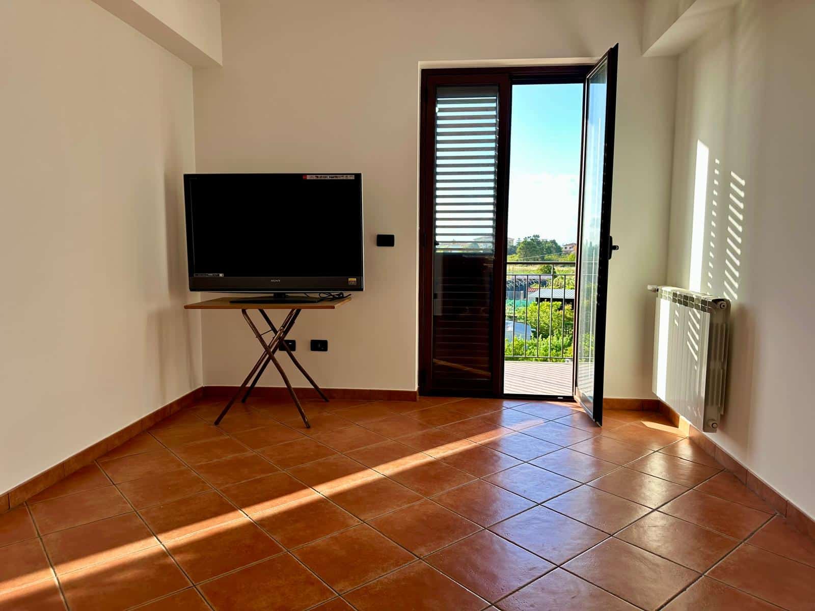 Il televisore in soggiorno con porta finestra aperta che dà su un balcone con vista verde, in una stanza luminosa e moderna.