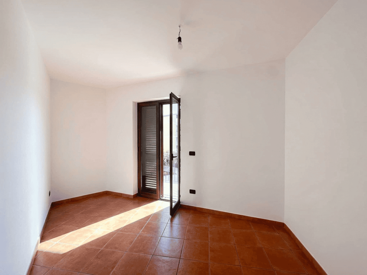 Appartamento vuoto con porta finestra che si apre su balcone, pavimento in ceramica rossa, pareti bianche, ideale per ristrutturazione o affitto.