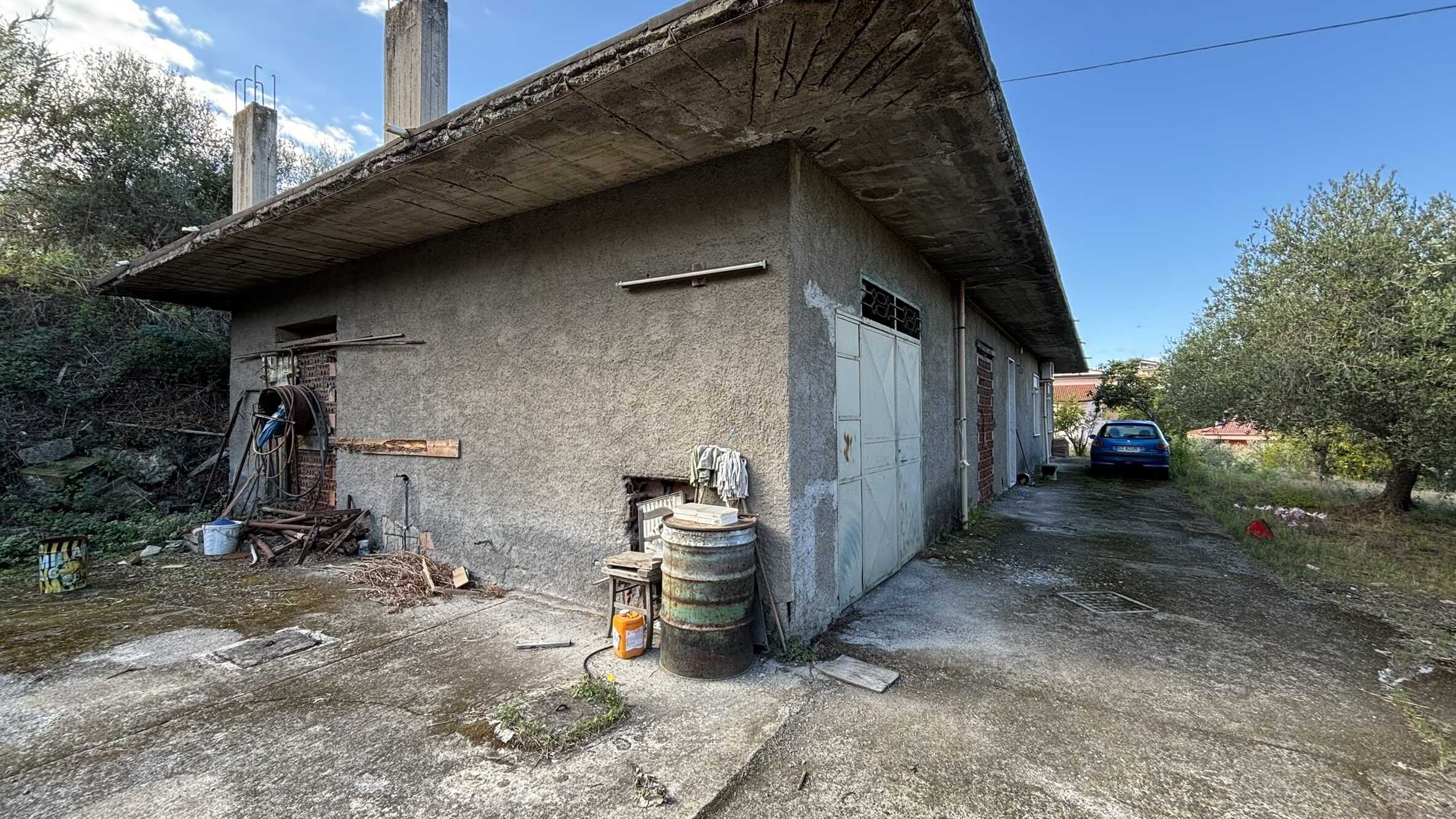 Vecchio capannone in penombra con pareti in cemento e tetto a spiovente, spazi di deposito e aree esterne con auto e vegetazione, destinato a ristrutturazione in zona rurale.