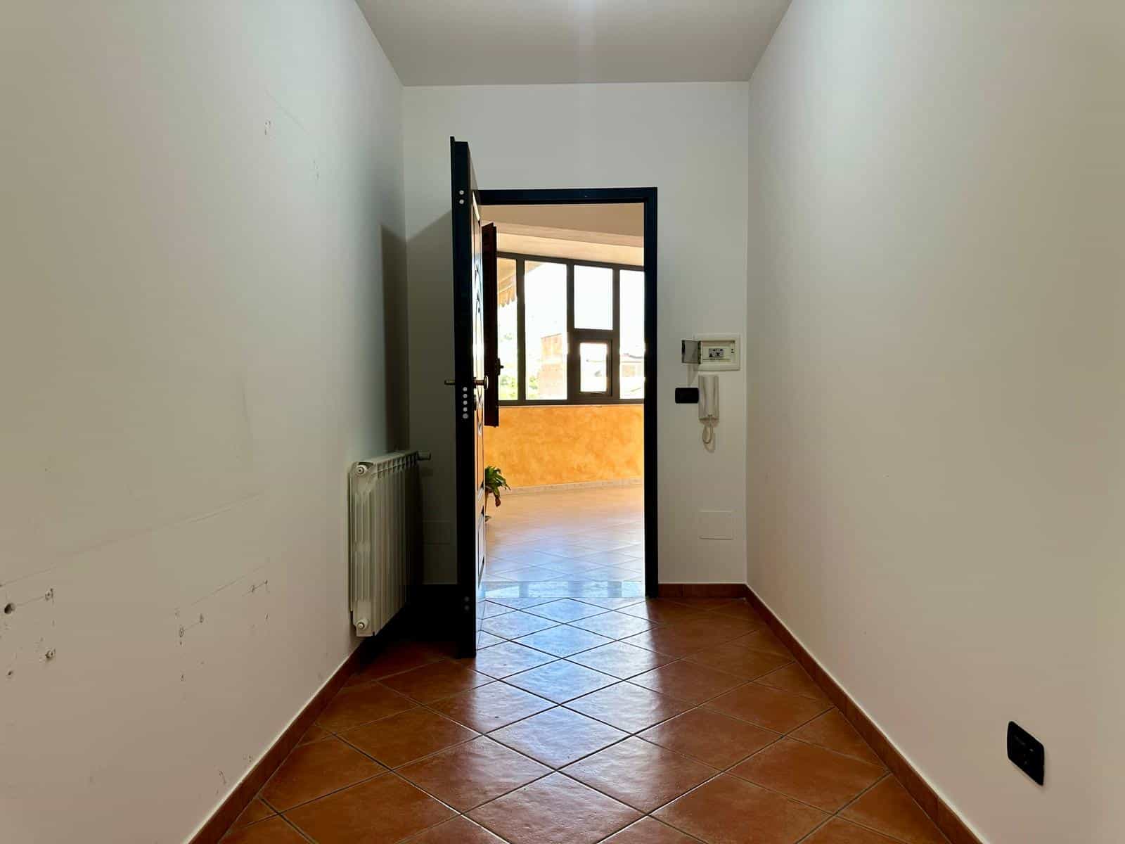 Ingresso di un appartamento con porta aperta che conduce a una stanza luminosa con grandi finestre e pavimento in piastrelle marroni.