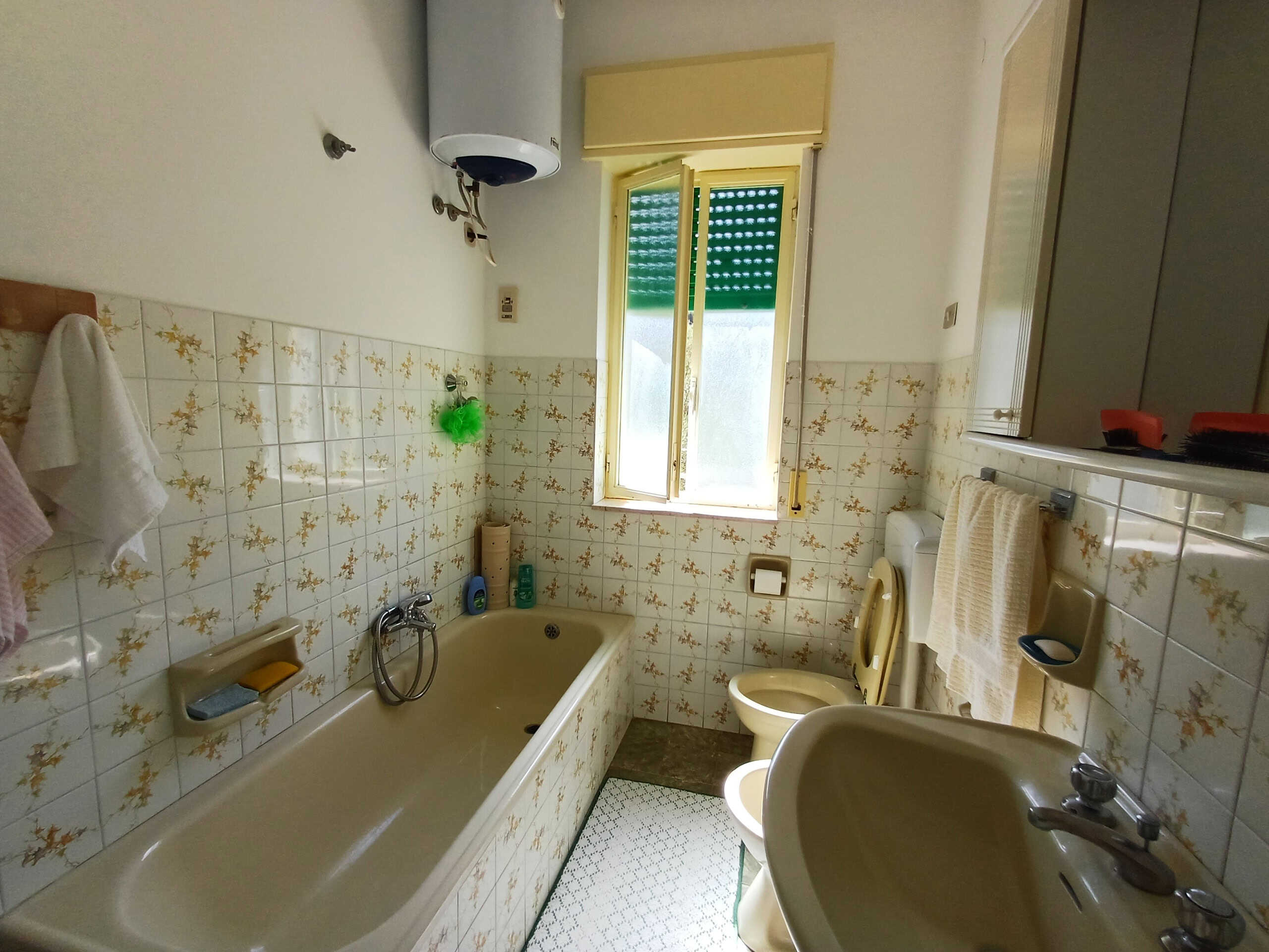 Doccia bagno con vasca in ceramica chiara, finestra aperta con zanzariera verde, pareti rivestite con piastrelle decorative a fiori, lavabo con piantana, tenda da bagno, generatore di calore, spazio sgombero per prodotti di igiene personale, illuminazione naturale, ambiente abitabile e funzionale.
