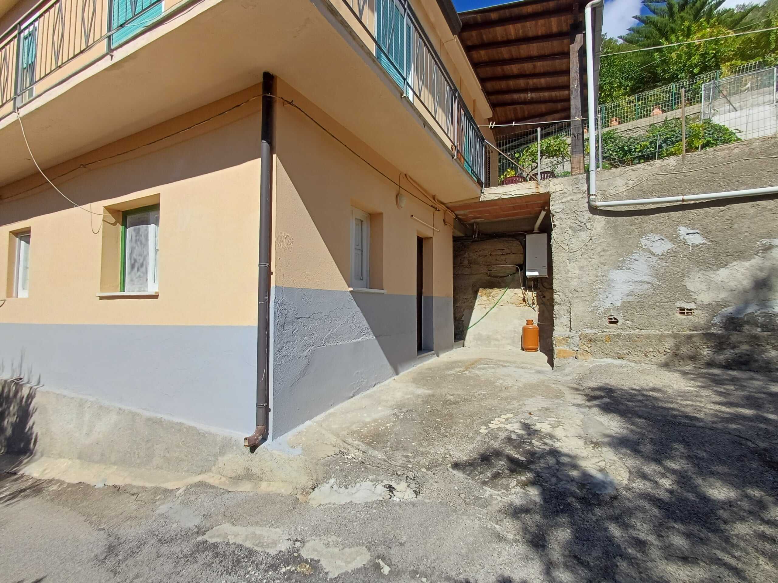 Finestra e ingresso di un appartamento su strada con pareti esterne verniciate in beige e grigio, vista da strada sterrata e asfalto, con elementi di ristrutturazione edilizia.