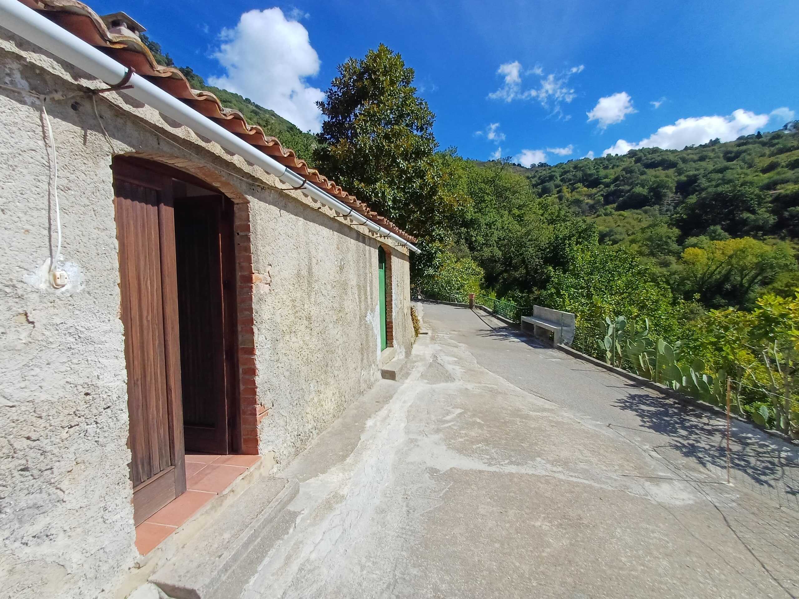 Vecchia casa di campagna in stile rustico con pareti in pietra, porta in legno e tetto in terracotta, immersa in un paesaggio collinare verde e soleggiato con cielo blu.