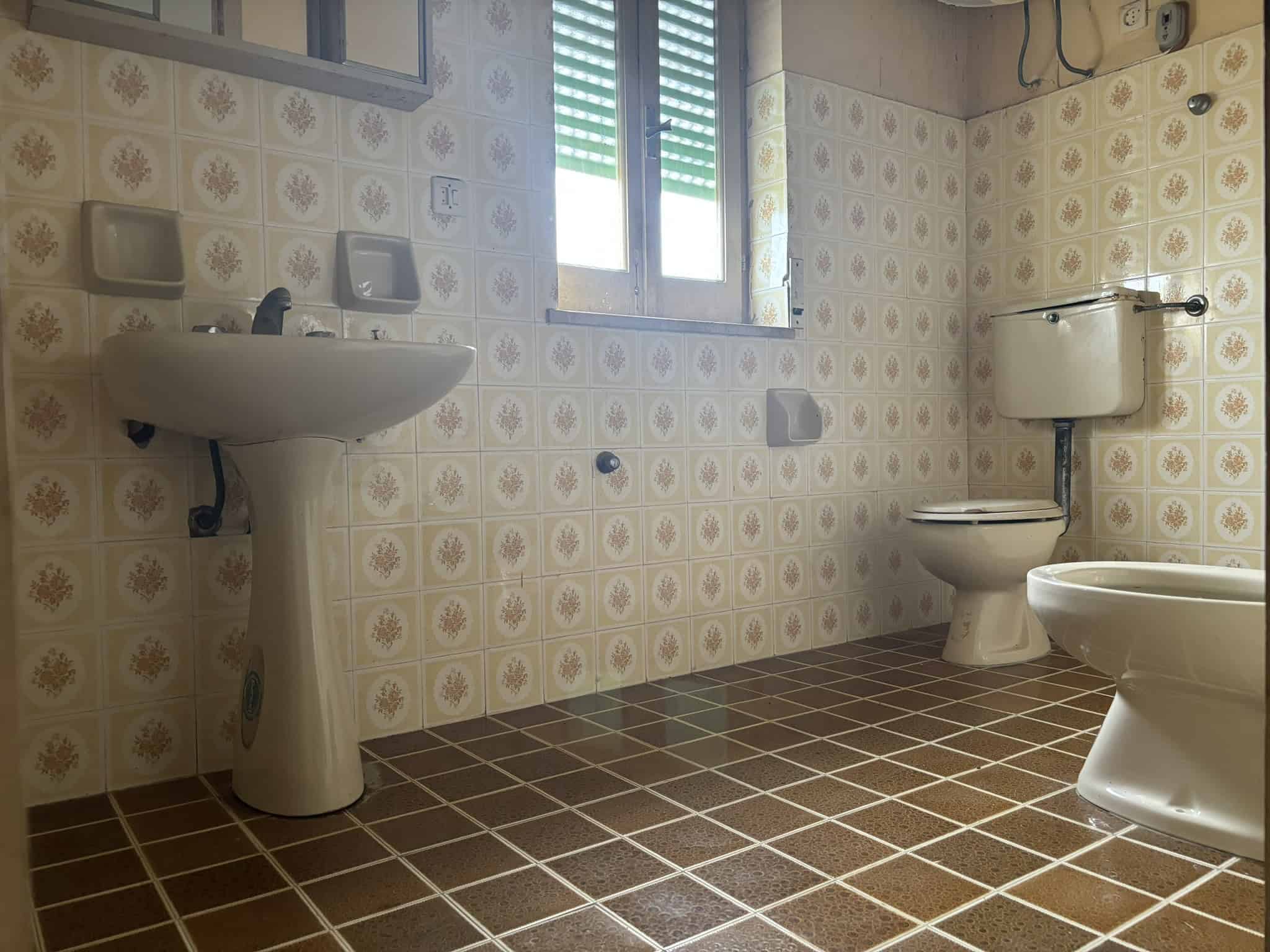 Lavabo e servizi igienici in bagno con pareti rivestite in piastrelle vintage beige e marroni, finestra con persiana, ambiente spazioso e funzionale, ideale per immobiliare e ristrutturazioni.
