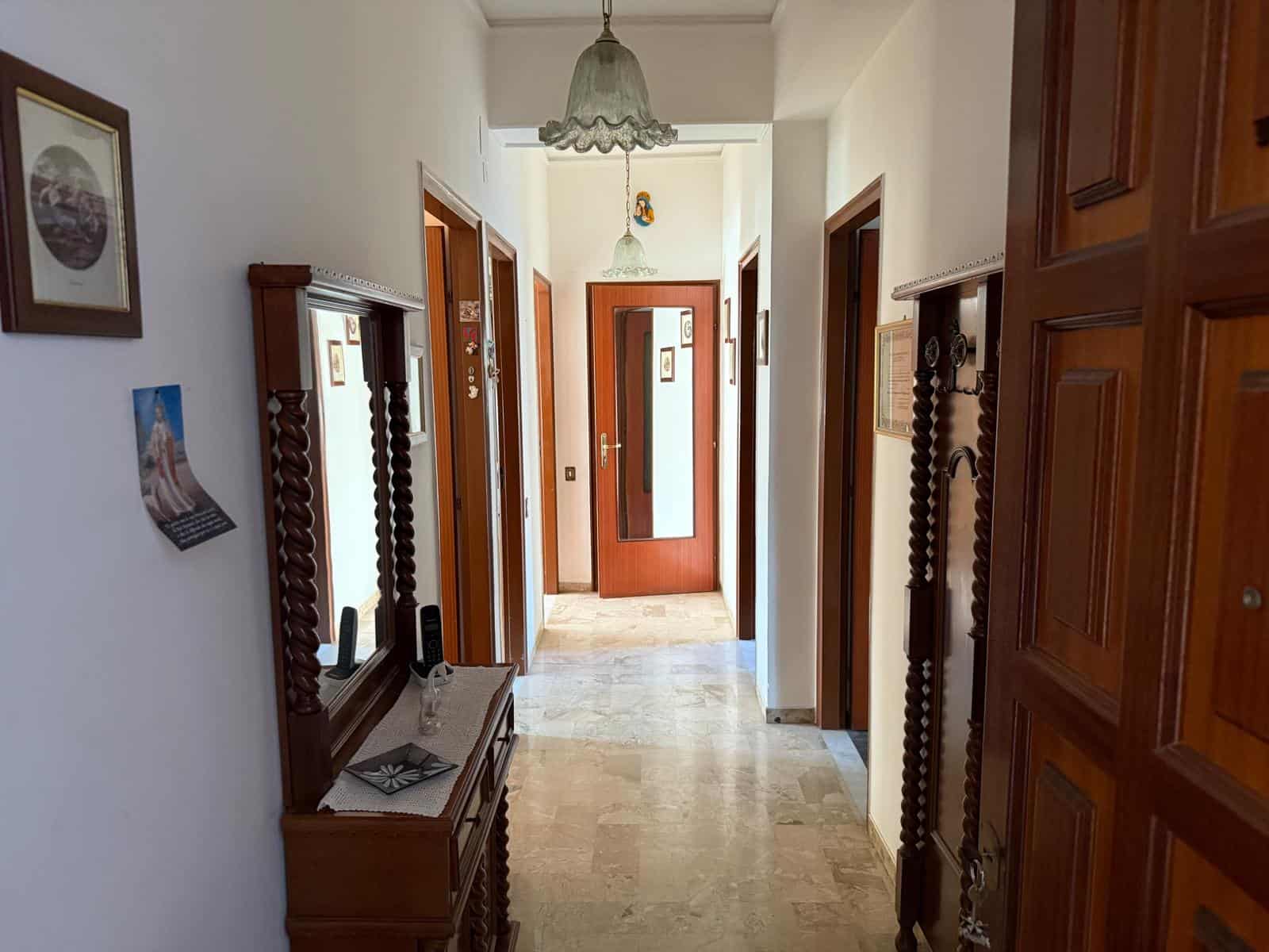 Porta d'ingresso in legno con finestra laterale, arredamento classico in corridoio, pareti decorate con quadri e specchi, pavimento in marmo beige, illuminazione naturale e lampadario vintage.