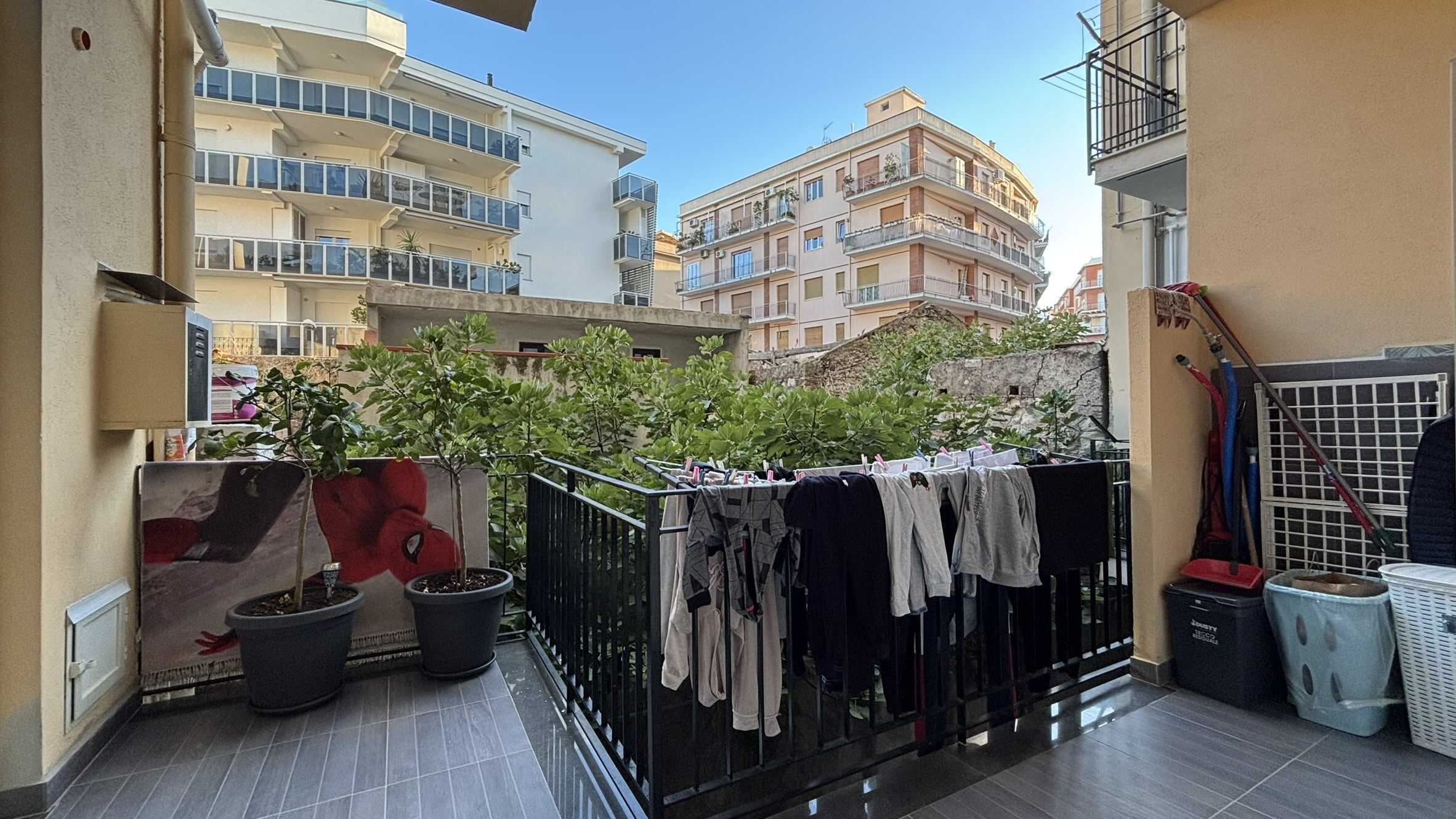 Balcone con piante, quadri, e abbigliamento steso, in un condominio moderno a Roma, vista su altri edifici residenziali, ideale per affitti turistici o investimento immobiliare.