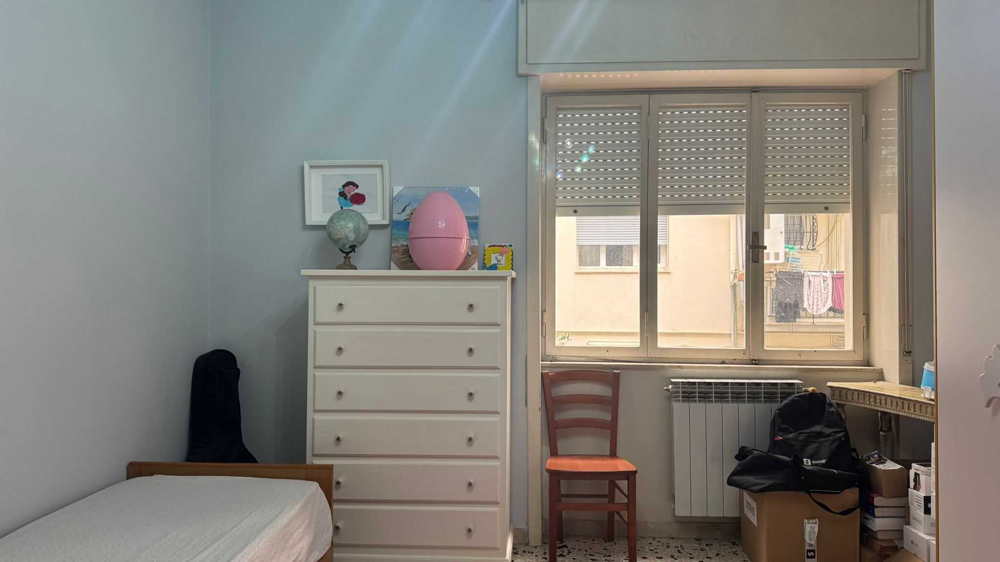 Letto singolo con arredamento semplice, finestra con persiane, stanza da letto arredata, oggetti vari sul comodino, atmosfera domestica e funzionale.
