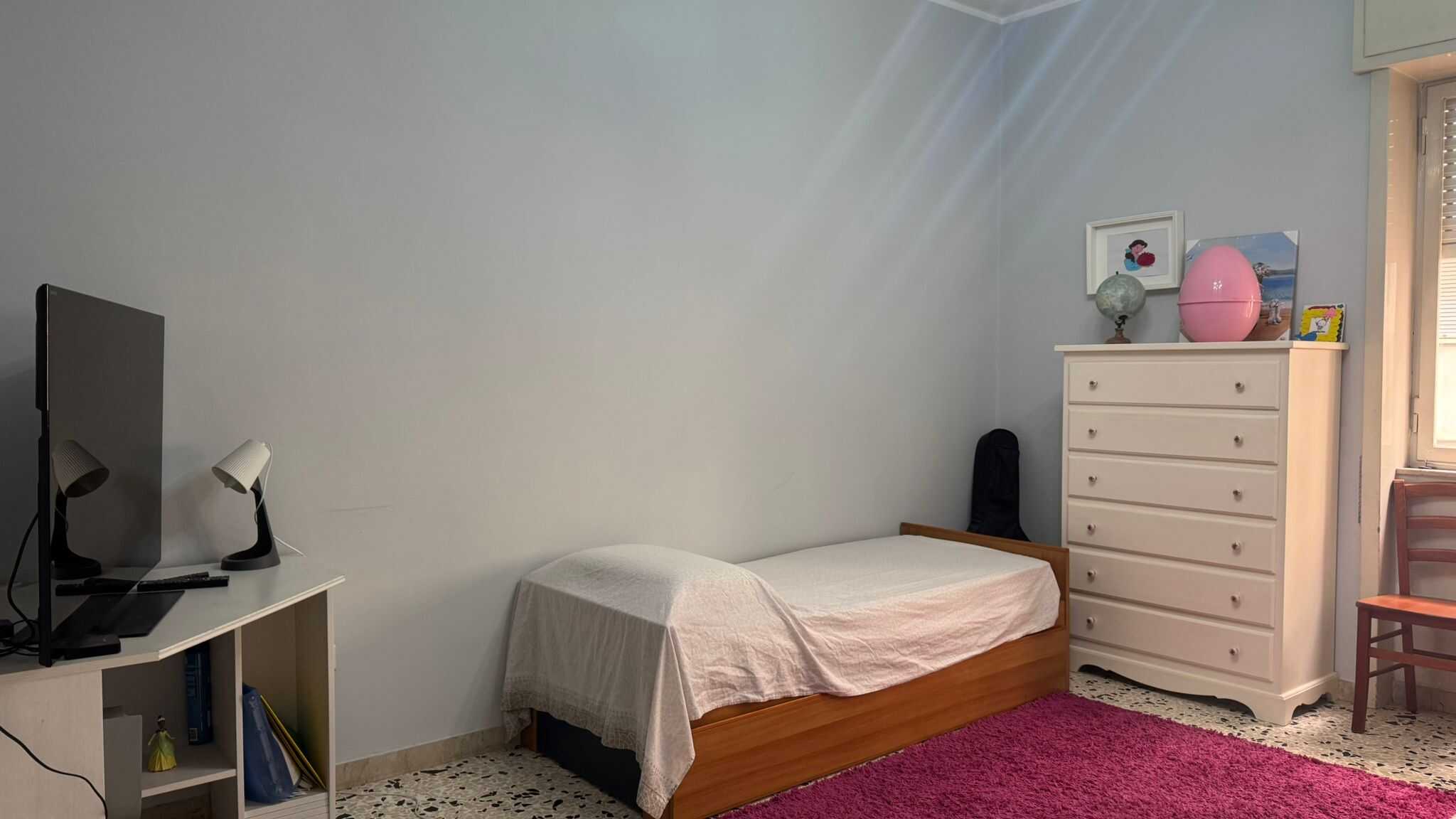 Letto singolo con copriletto bianco, armadio bianco con decorazioni colorate sulla parte superiore, piccola scrivania con monitor e lampade da tavolo, tappeto rosso, finestra con persiane, scena di una camera da letto in stile semplice e accogliente.