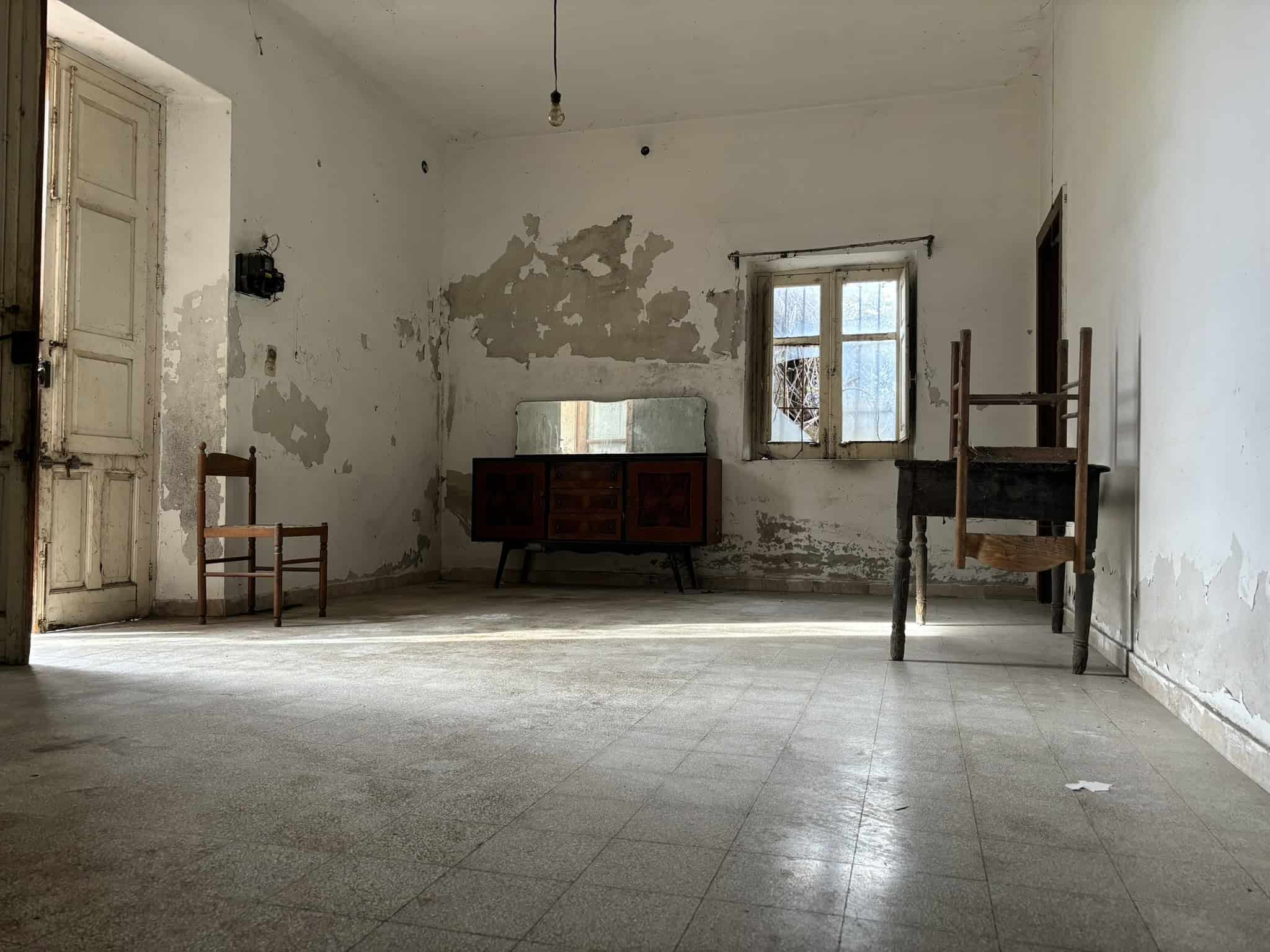 Vecchio soggiorno in condizioni di rovina, con pareti danneggiate e arredi vintage in un ambiente abbandonato. La stanza presenta una finestra rotta e uno stile rustico e trasandato.