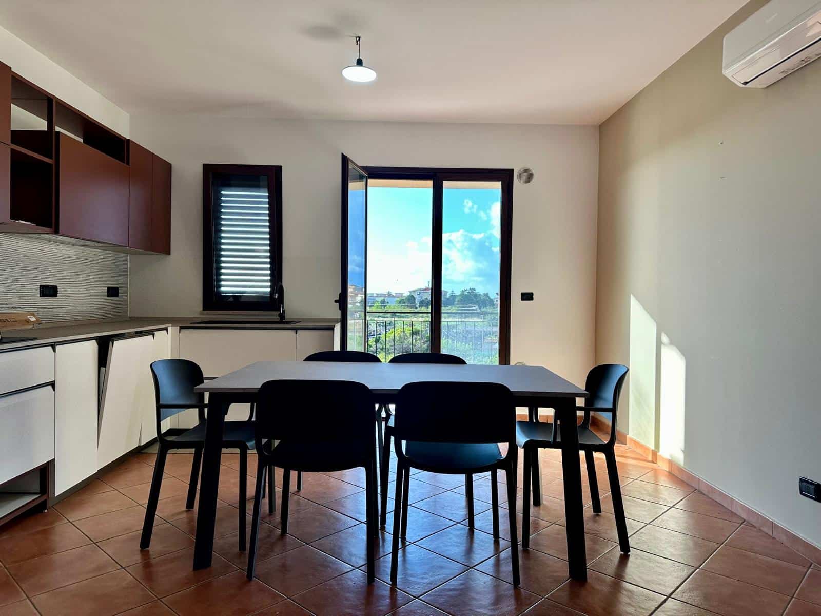 Interno di una sala da pranzo moderna con tavolo nero, sedie nere, cucina a vista e grande finestra con vista panoramica sulla città e il cielo blu.