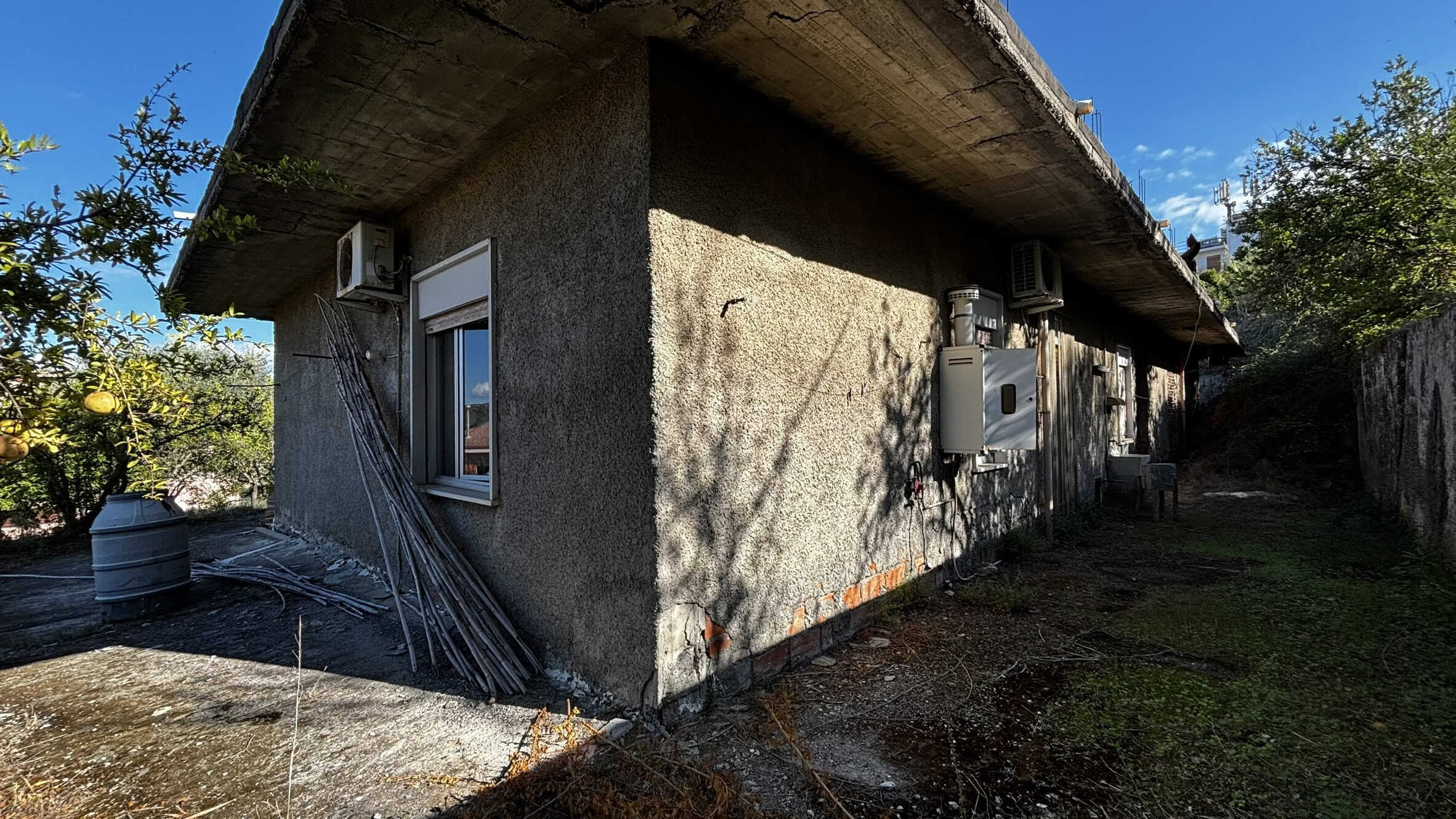 Vecchia casa da ristrutturare con pareti in cemento e finestre a villino, esterno con giardinetto e alberi da frutto, in zona tranquilla e verde.