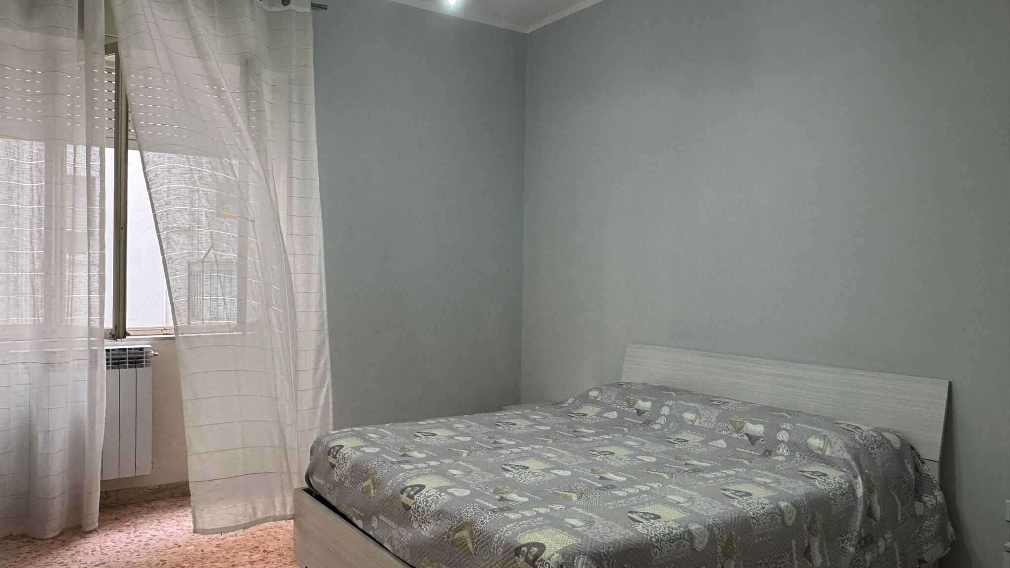 Letto matrimoniale in camera con pareti azzurro chiaro e finestra con tende leggere, ambientazione accogliente e luminosa.