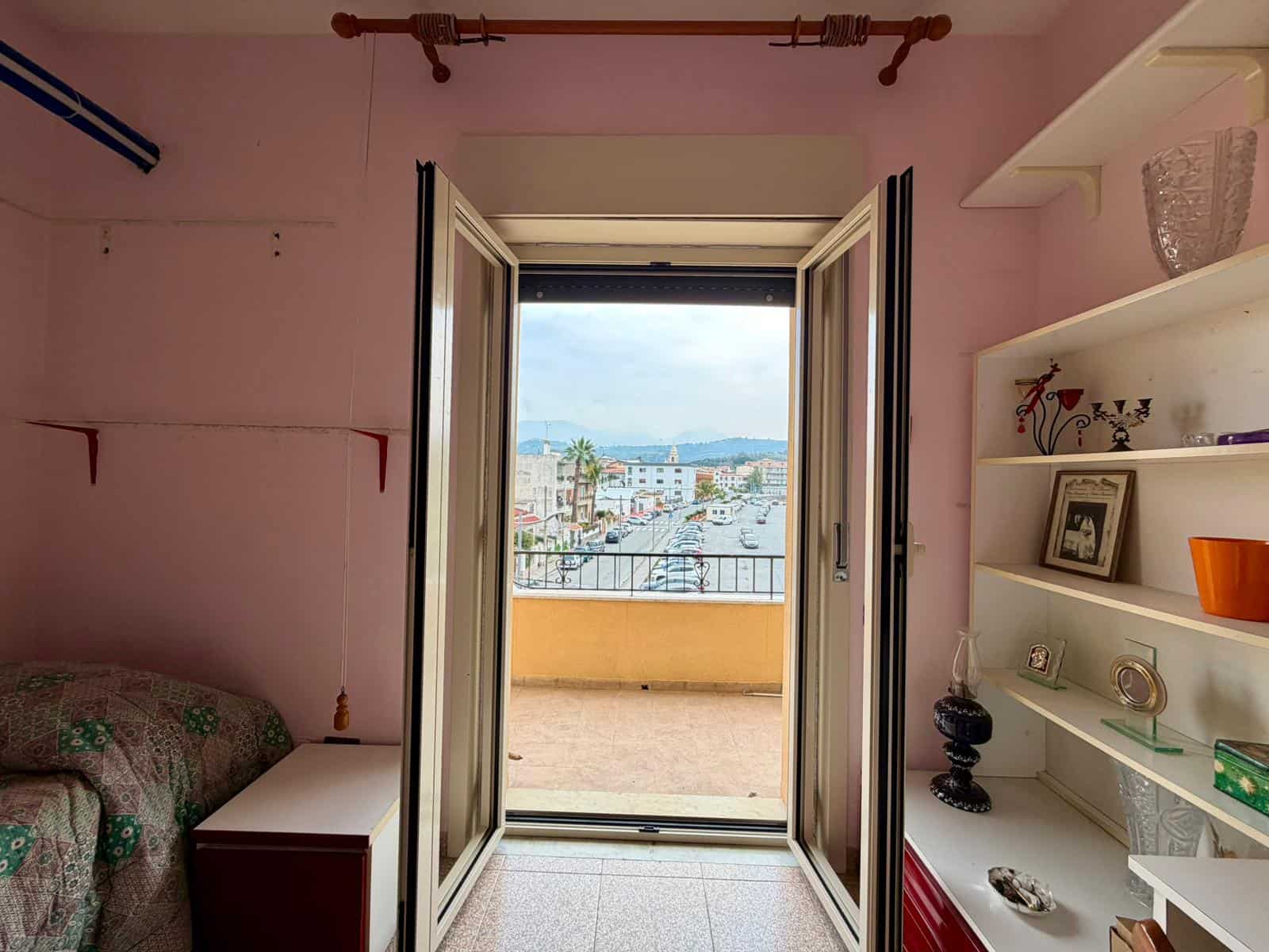 Finestra aperta con vista sul porto e sulla città, illuminazione naturale in una stanza con pareti rosa e scaffali decorativi.