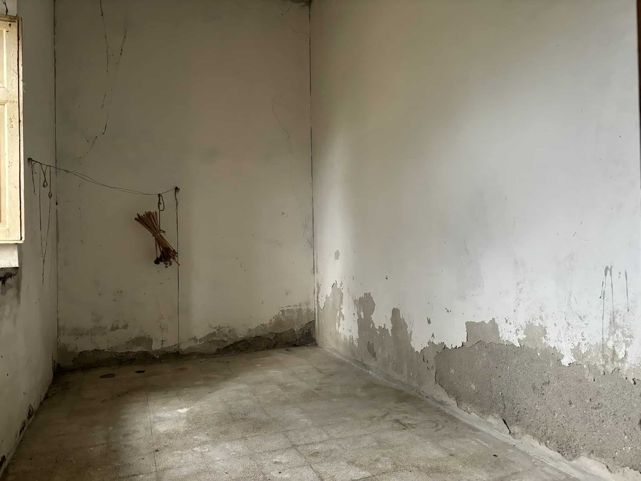 Vecchio locale da ristrutturare, pareti con muffa e crepe, pavimento in ceramica usurato, ambiente di scarsa luminosità, ideale per progetto di ristrutturazione per appartamento o casa privata.