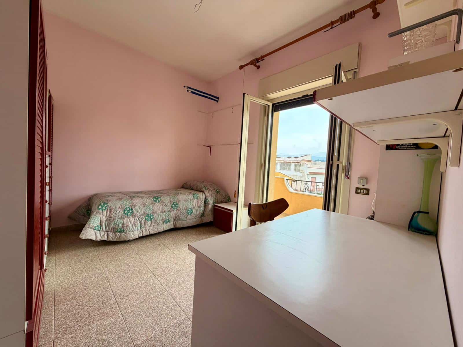 Camera da letto luminosa con pareti rosa, letto singolo e balcone con vista panoramica sulla città, ideale per affitti brevi o soggiorni rilassanti a NextCasa.
