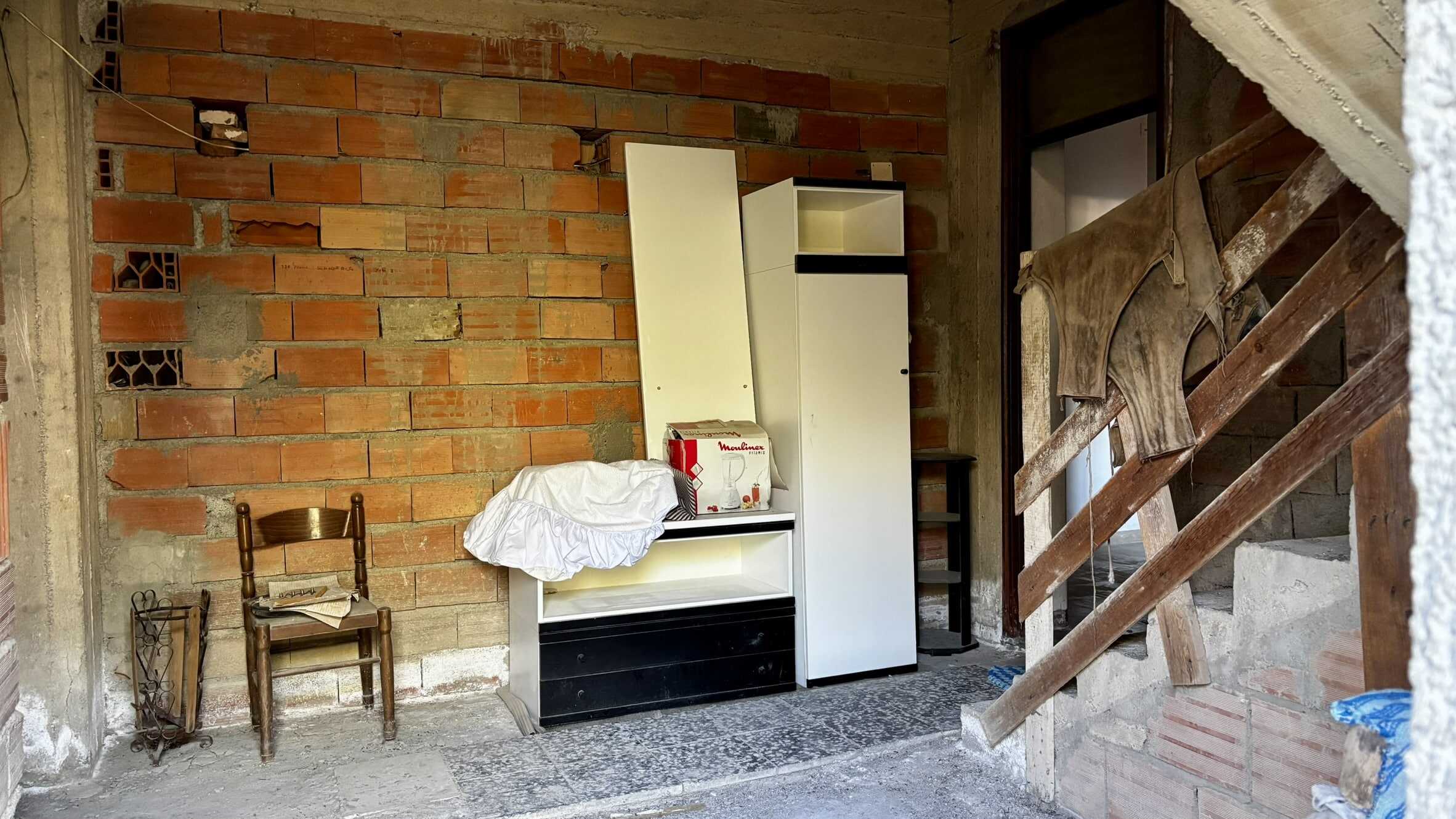 Vecchia casa in costruzione con pareti in mattoni rossi e arredi essenziali, scaffali e sedie in legno, ambiente rustico e ancora in fase di ristrutturazione.