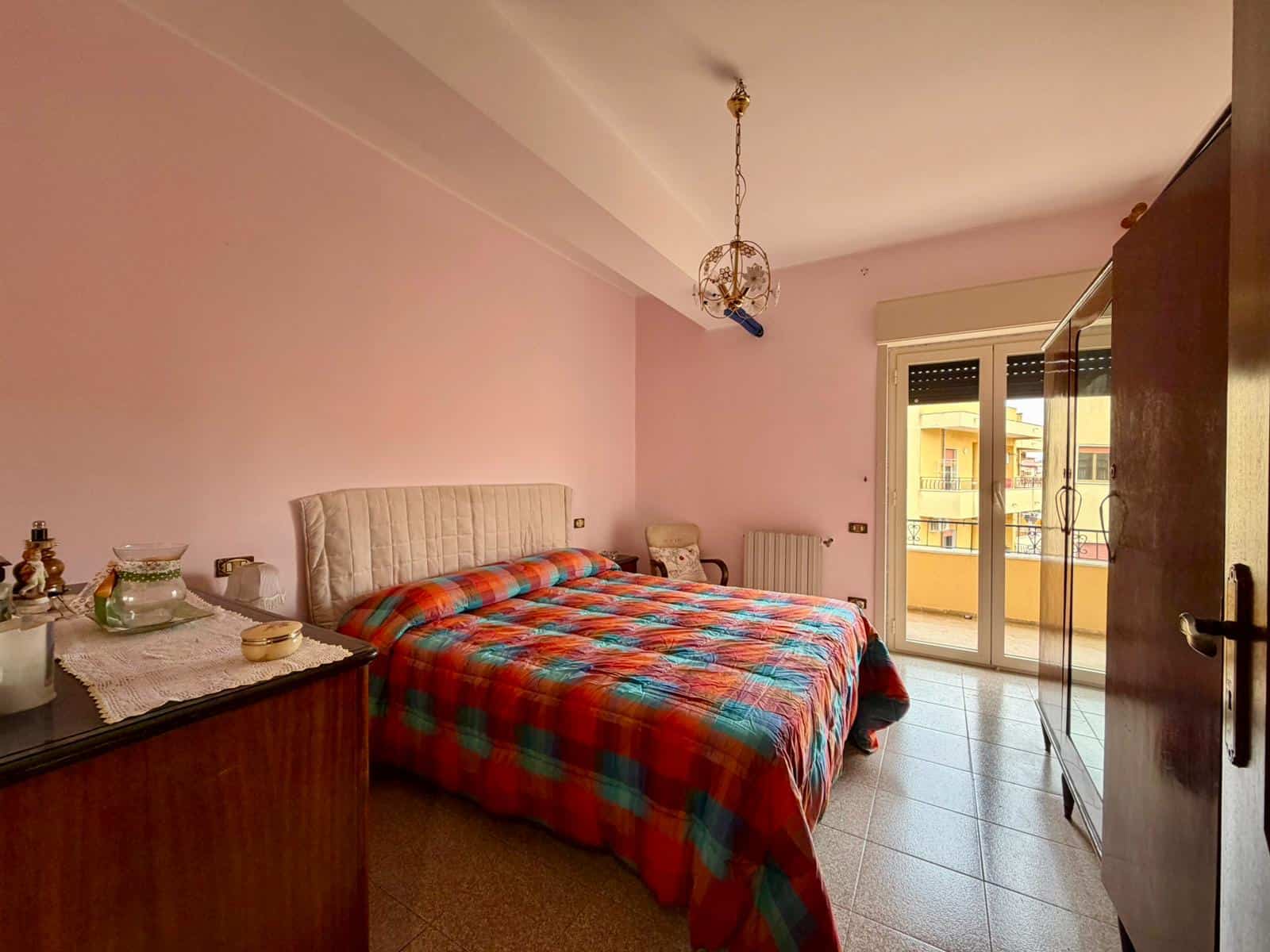 Ampio soggiorno con pareti rosa, letto matrimoniale con coperta a quadri colorati, armadio grande e balcone con vista sulla città, ideale per affitti residenziali o turistici.