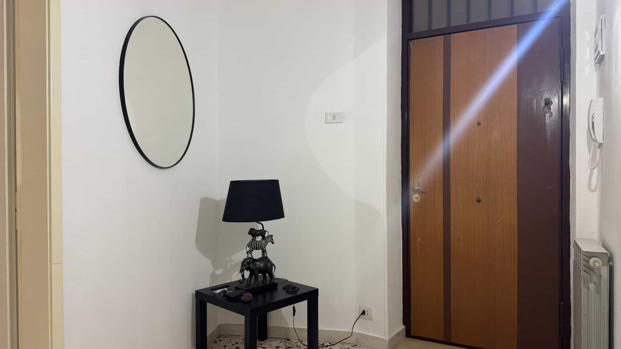 Porta d'ingresso moderna in legno con dettaglio nero, specchio decorativo rotondo e specchiera, lampada stilizzata con zebra, interruttori e telefono fisso, ambiente elegante e funzionale.