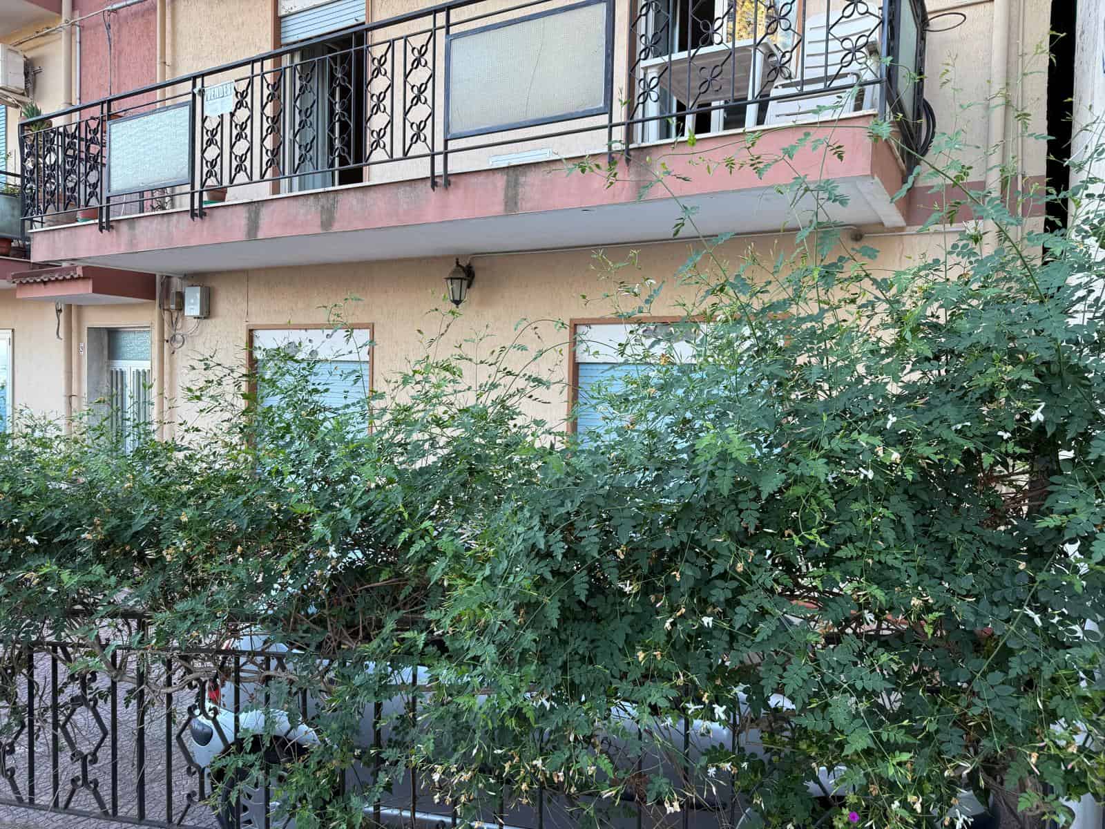 Balcone con ringhiera in ferro battuto e piante verdi in primo piano, appartamento in vendita con balcone e finestre con tende e porte a vetri, edificio residenziale con caratteristiche tipiche per il settore immobiliare.