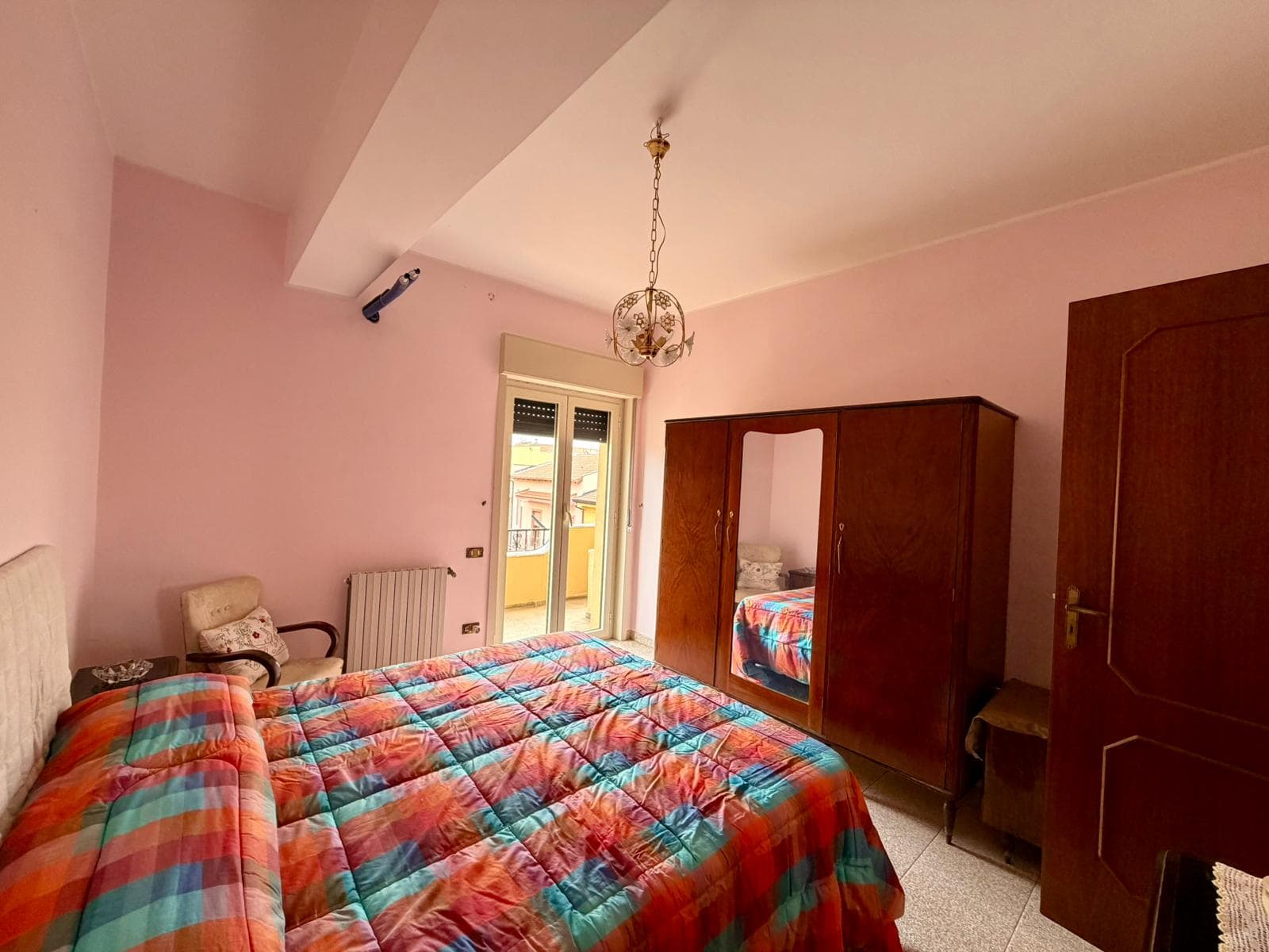 Riproduzione di una camera da letto con pareti rosa chiaro, letto matrimoniale con copriletto a quadri colorati, armadio in legno con specchio, porta in legno e finestra con vista su un balcone. Atmos.
