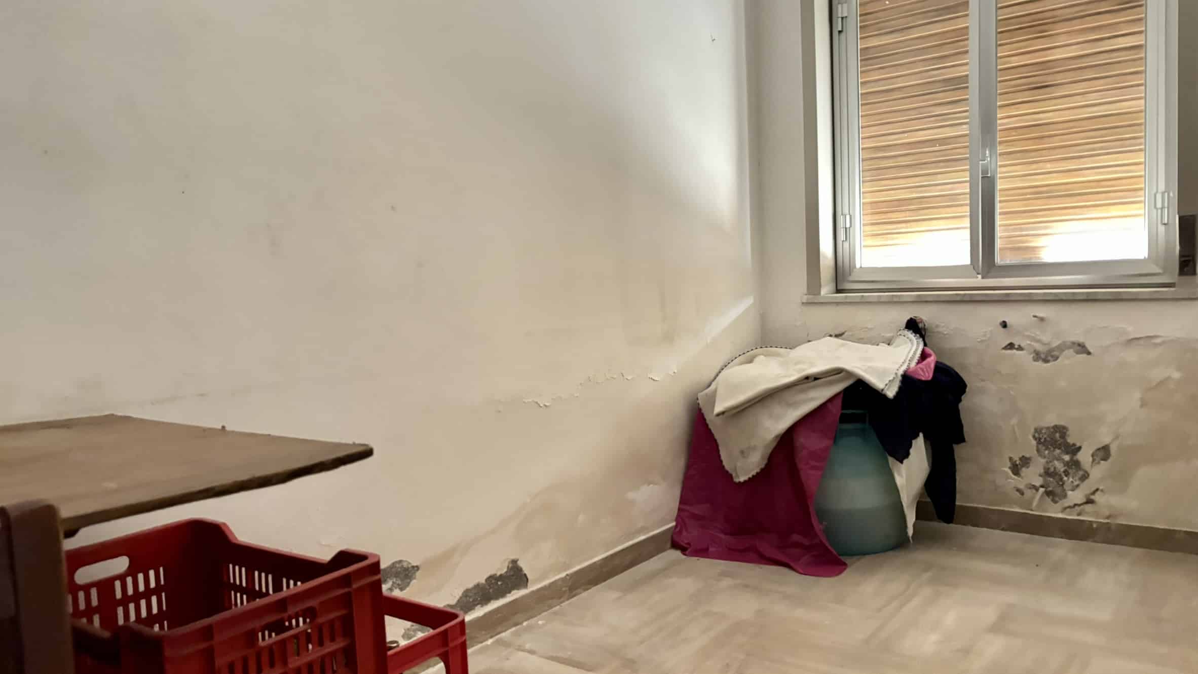 Vecchia parete con segni di umidità e pittura scrostata in una stanza da ristrutturare, di fronte a una finestra con tapparelle di legno, elementi tipici di immobili da riqualificare in zona residenziale.