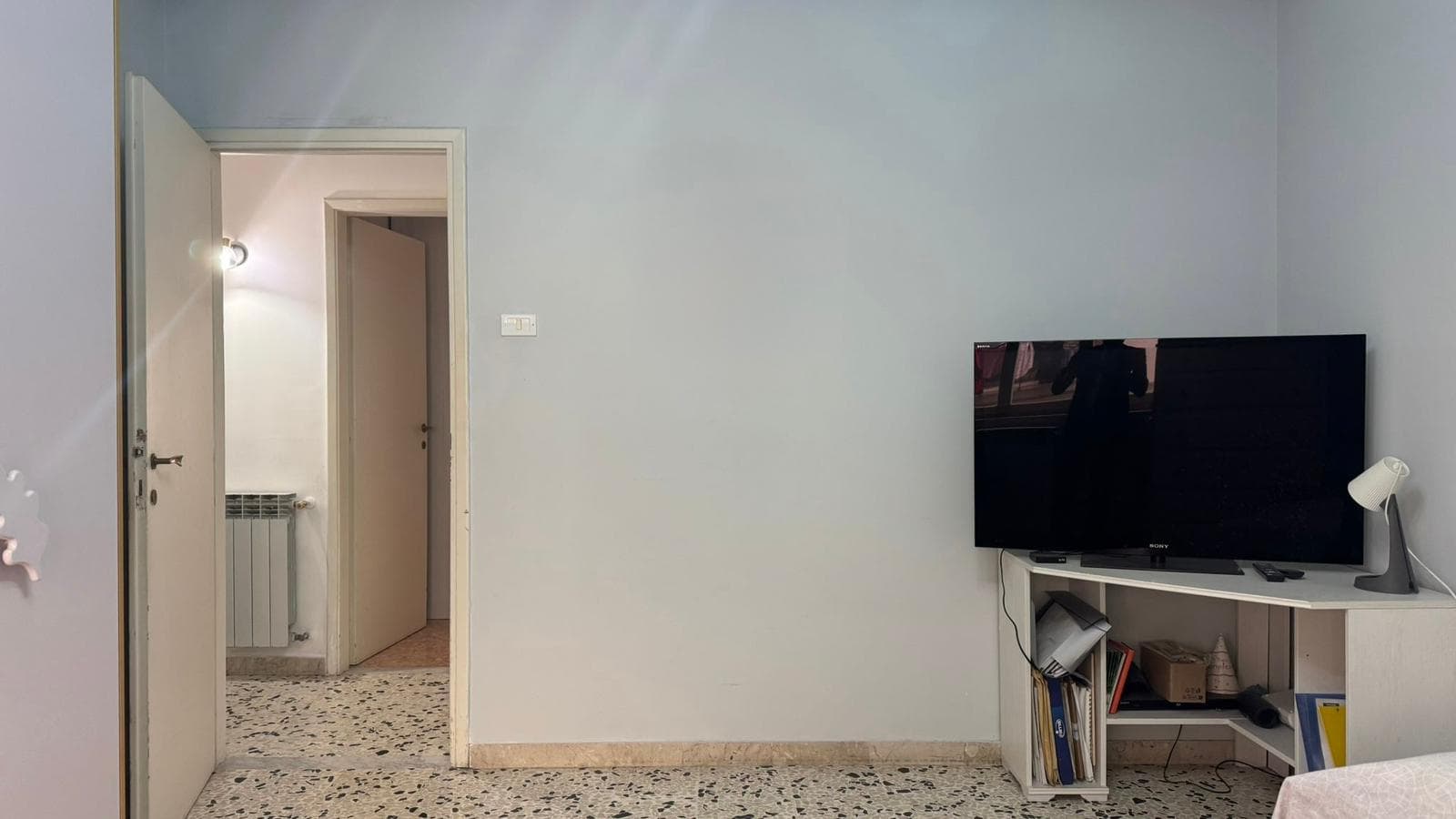 Televisore LED in soggiorno con mobile porta TV, lampada da tavolo e libri, pareti tinteggiate di colore chiaro, ingresso con porta aperta, ambiente domestico accogliente e funzionale.