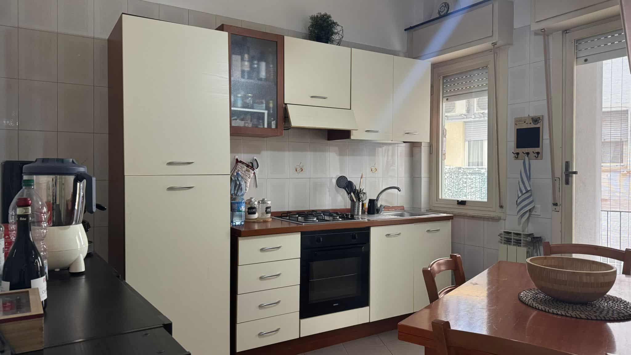 Cucina abitabile con armadi a ante bianche e dettagli in legno, dotata di forno, piano cottura e lavabo, vicino a finestra luminosa che permette l'ingresso di luce naturale, ambiente funzionale e accogliente.