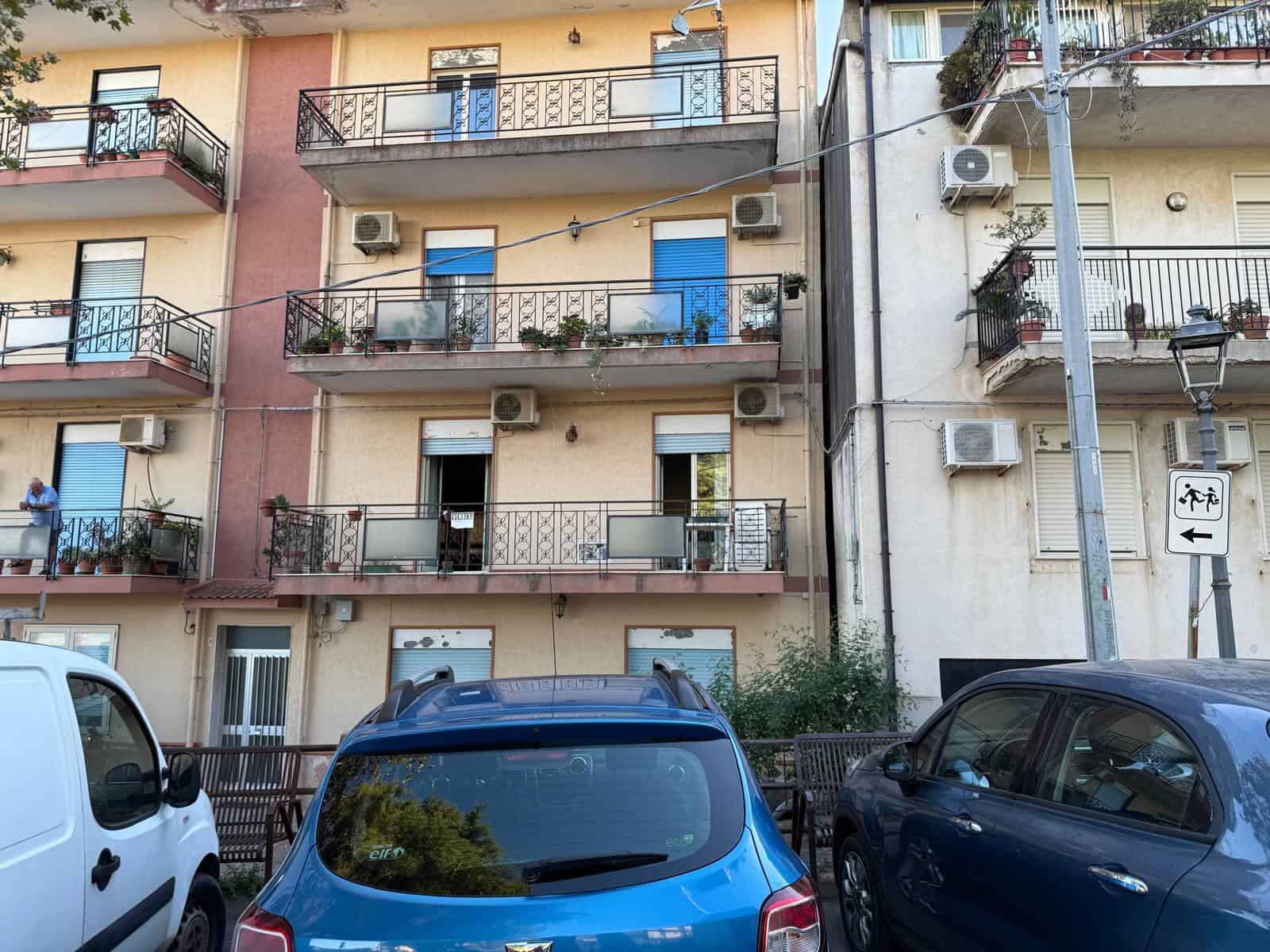 Immagine di un edificio residenziale con facciata colorata, balconi con piante e condizionatori, zona parcheggio con auto e un lampione, in un quartiere urbano.