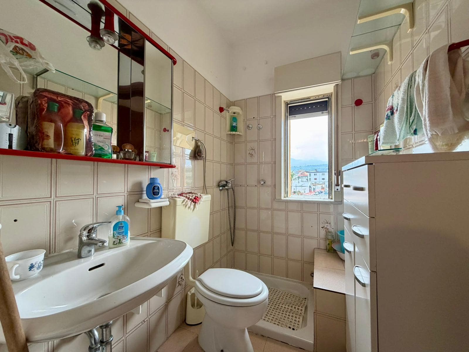 Lavabo e servizi igienici in un bagno compatto con finestra, piastrelle beige, armadietto bianco e accessori essenziali, ideale per spazi piccoli e funzionali.