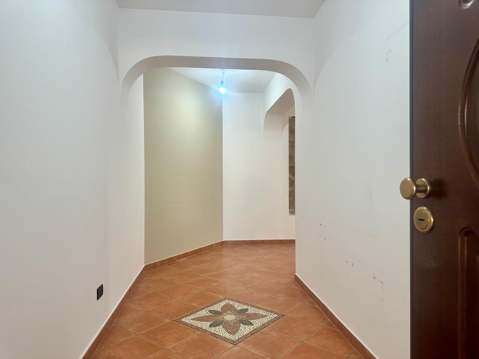 Ampia entrata di casa con pareti bianche, pavimento in piastrelle di ceramica con motivo decorativo centrale, e arco che conduce a un corridoio luminoso.