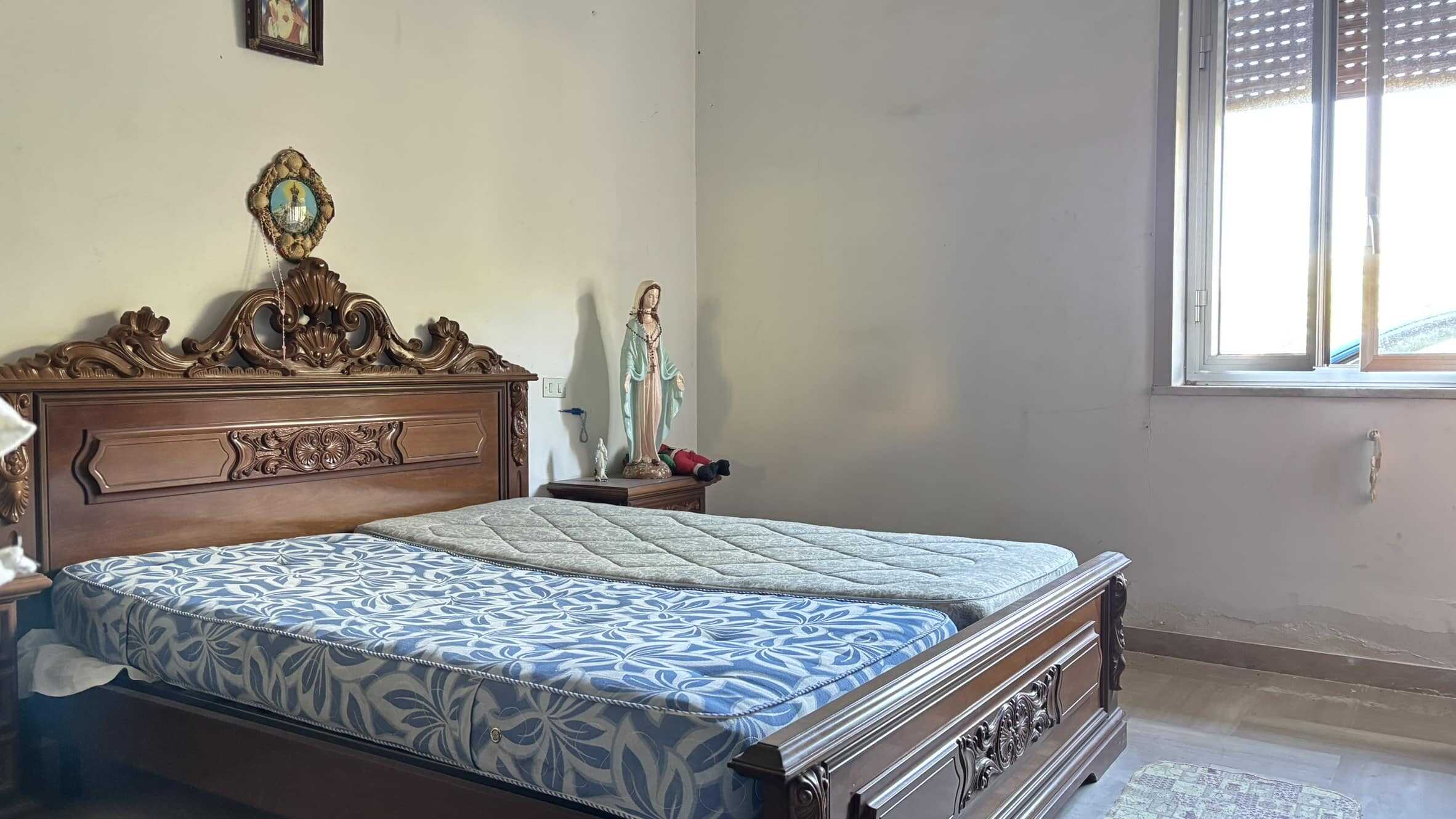 Letto matrimoniale in stile classico con testata intarsiata, camera da letto luminosa con finestra, arredi tradizionali e decorazioni religiose, ideale per case in vendita o affitto a NextCasa.