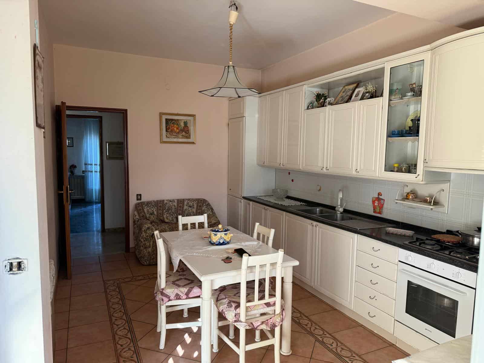Autoreggente cucina abitabile con mobili bianchi, tavolo da pranzo con sedie, elettrodomestici e decorazioni, ambiente luminoso e accogliente, ideale per famiglie e coppie.