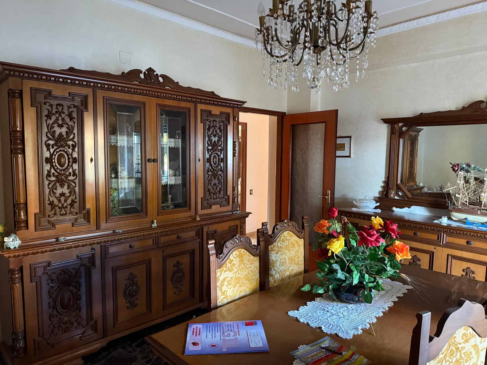 Elegante sala da pranzo con mobili in legno intarsiato, grande specchio e lampadario di cristallo, decorata con vaso di fiori colorati, perfetta per ambienti classici e raffinati.