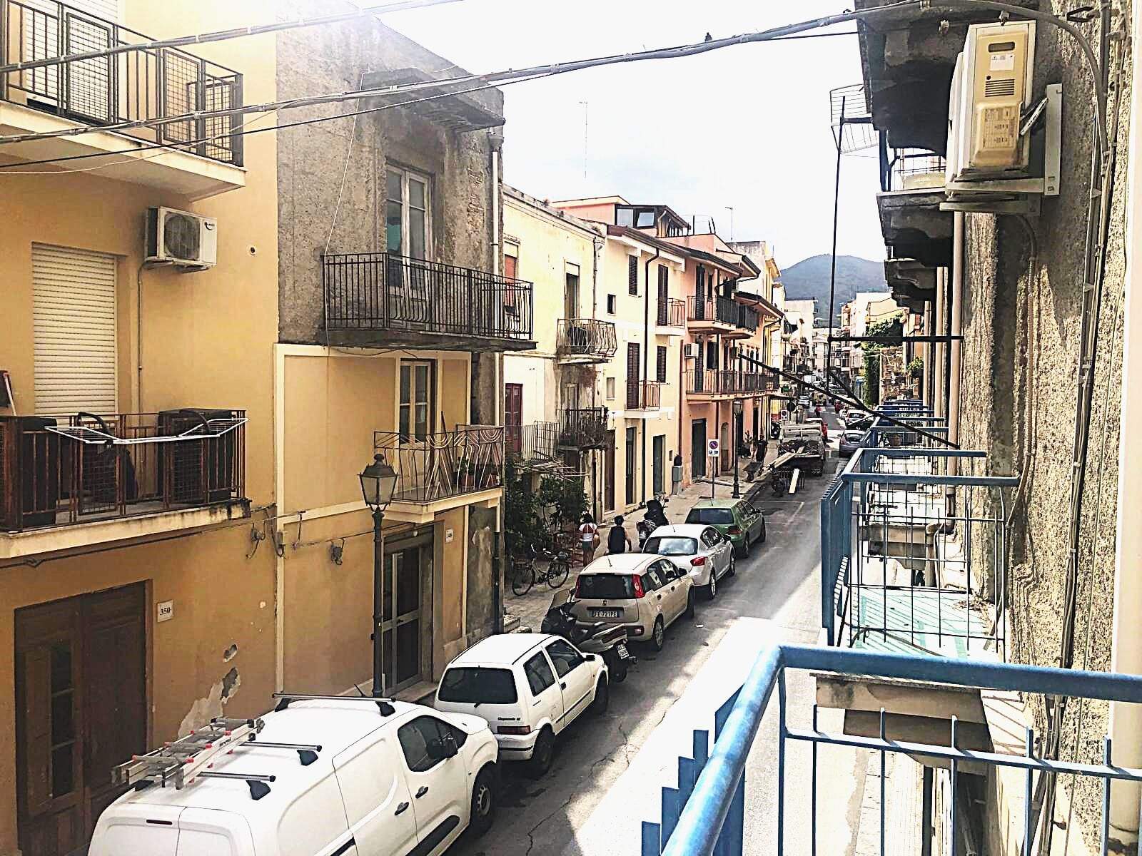 Finestra su un varco cittadino con edifici storici e auto parcheggiate, vista da un balcone in zona urbana di Napoli, con montagna sullo sfondo, su un giorno soleggiato.