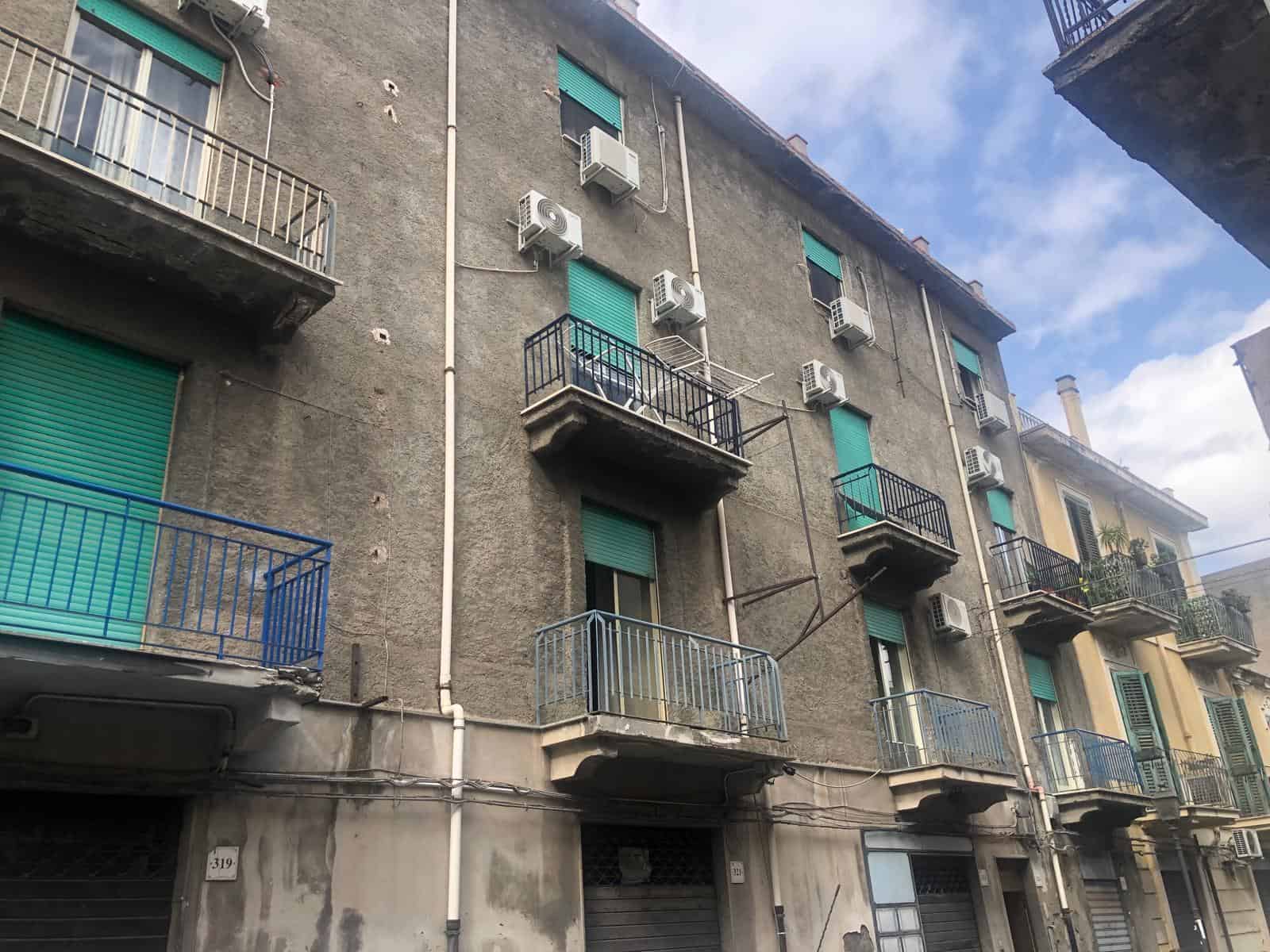 Balconi con rivestimento in metallo e climatizzatori su edificio residenziale nel centro urbano, con dettaglio di finestre con tapparelle verdi e facciata in pietra grigia, in un ambiente urbano italiano.