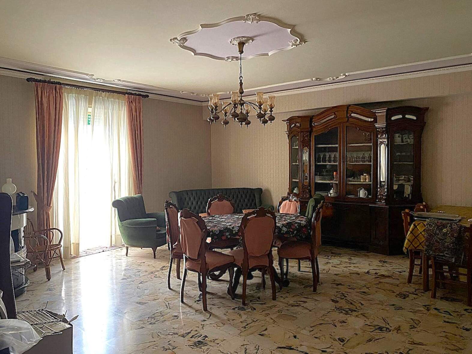 IMG-20250917-WA0009 Soggiorno elegante con tavolo da pranzo e sedie in stile classico, grande vetrina in legno e divano; ambienti luminosi con tende colorate e soffitto decorato, arredamento in stile tradizionale.
