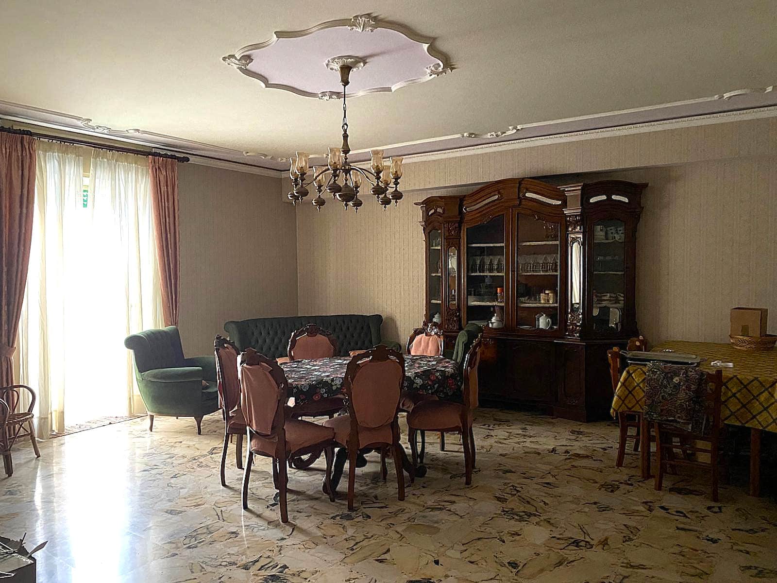 Antico soggiorno con tavolo da pranzo, divani e vetrina in legno scuro, decorazioni classiche, parquet in marmo, ampie finestre con tende color pesca, illuminazione a lampadario in stile vintage.