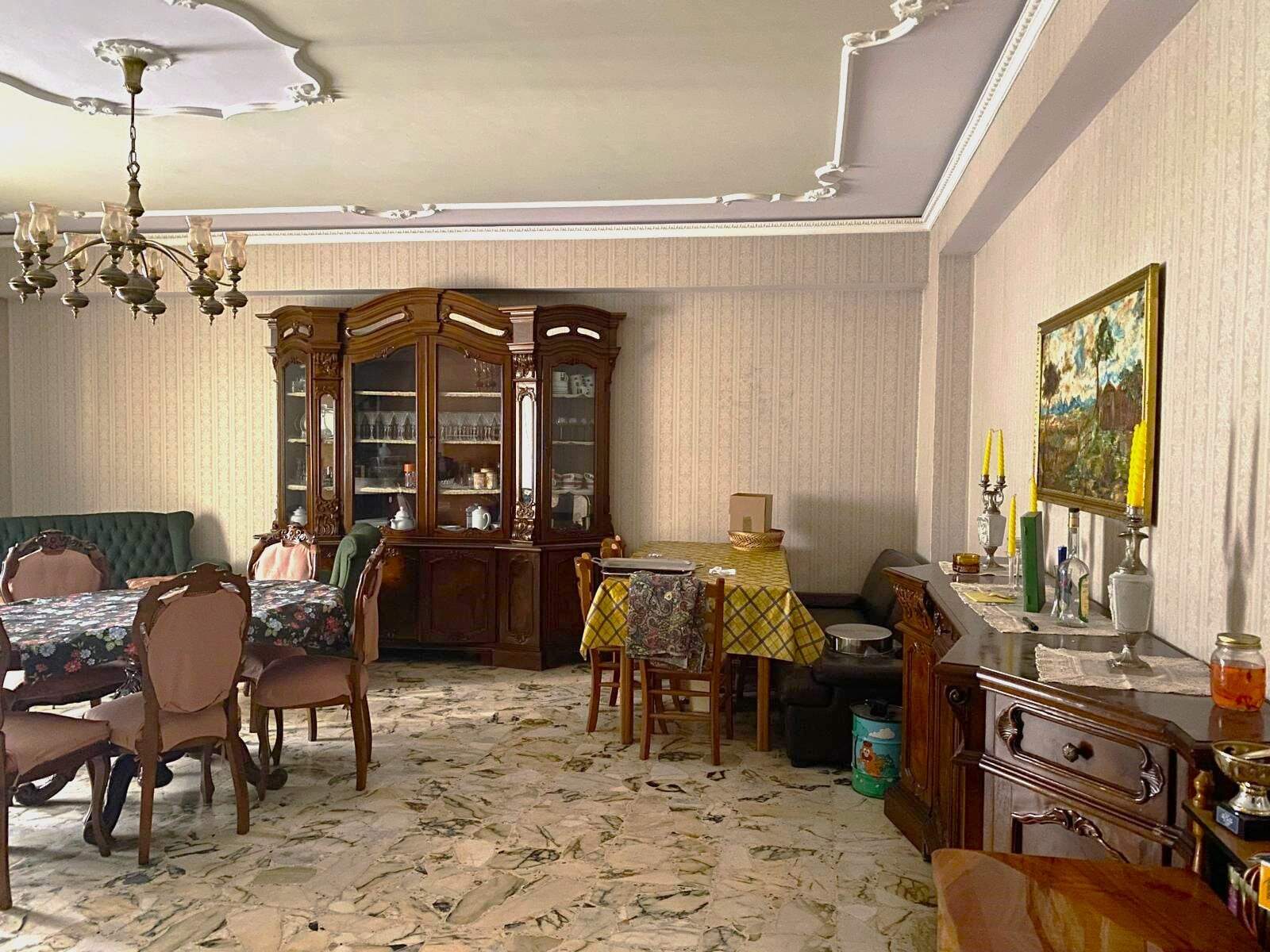 Antico arredamento di soggiorno con mobili in legno scuro, tavolo con tovaglia floreale, credenze e poltrone imbottite in stile classico, decorazioni vintage, pareti a righe e pavimento in marmo chiaro.