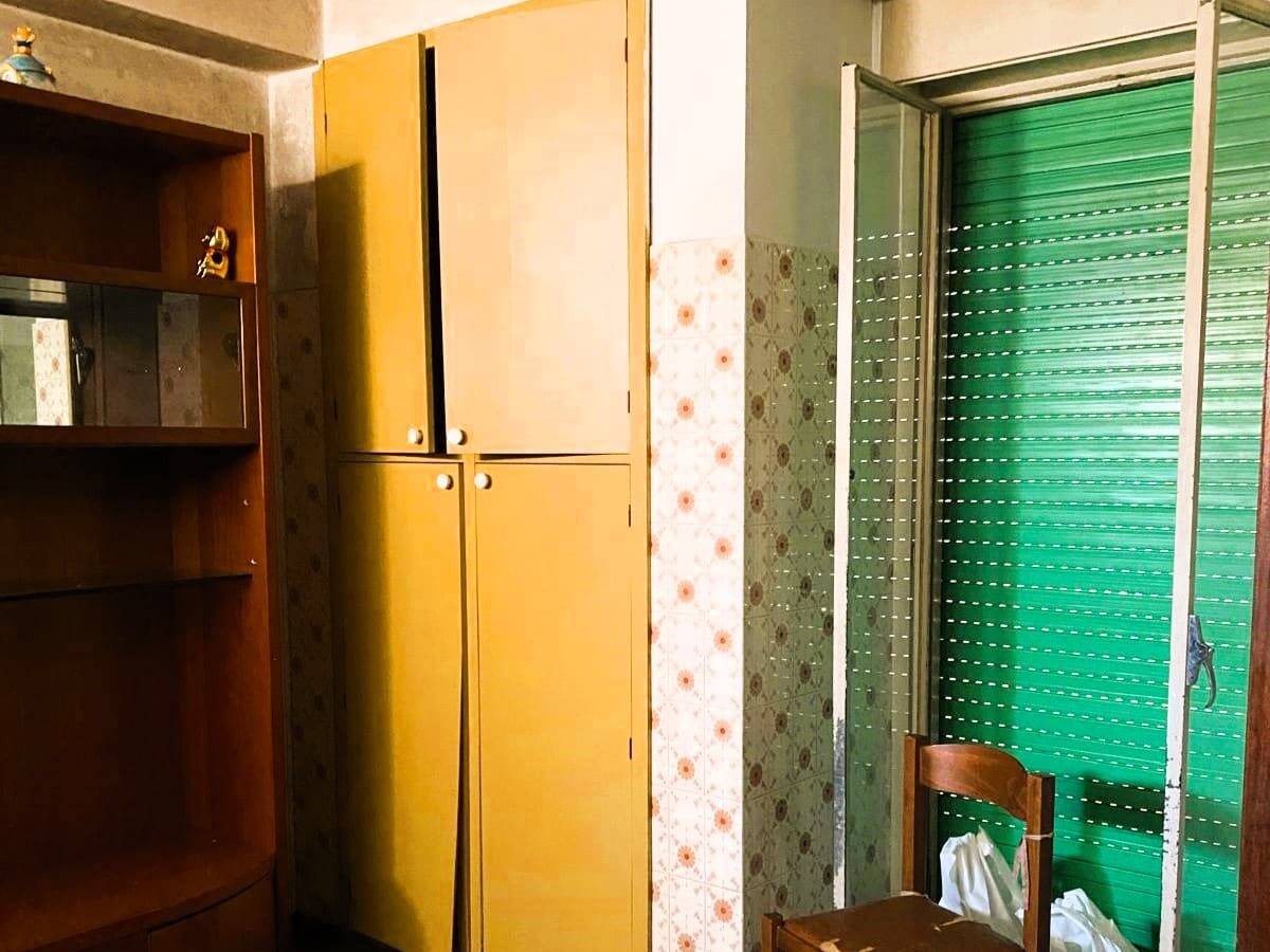 Armadio giallo e scaffale in legno per camera da letto, con porta finestra con avvolgibile in color verde, interni case vacanza NextCasa, arredamento funzionale e pratico, atmosfera casalinga e accogliente.