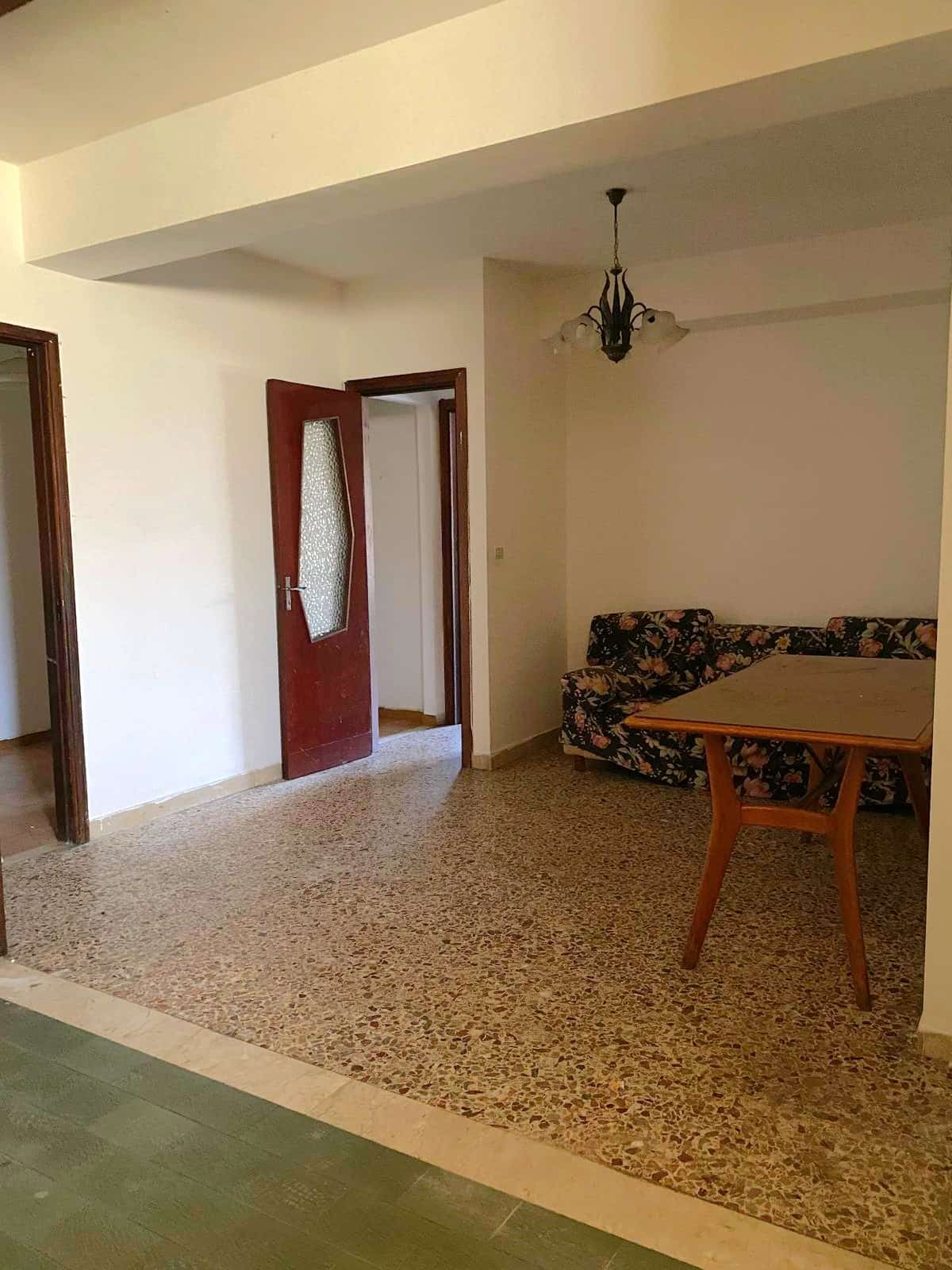 Soggiorno con arredo classico, divano con rivestimento floreale e tavolo in legno, illuminazione vintage, pavimento in graniglia e porte in legno, ambiente luminoso e accogliente.