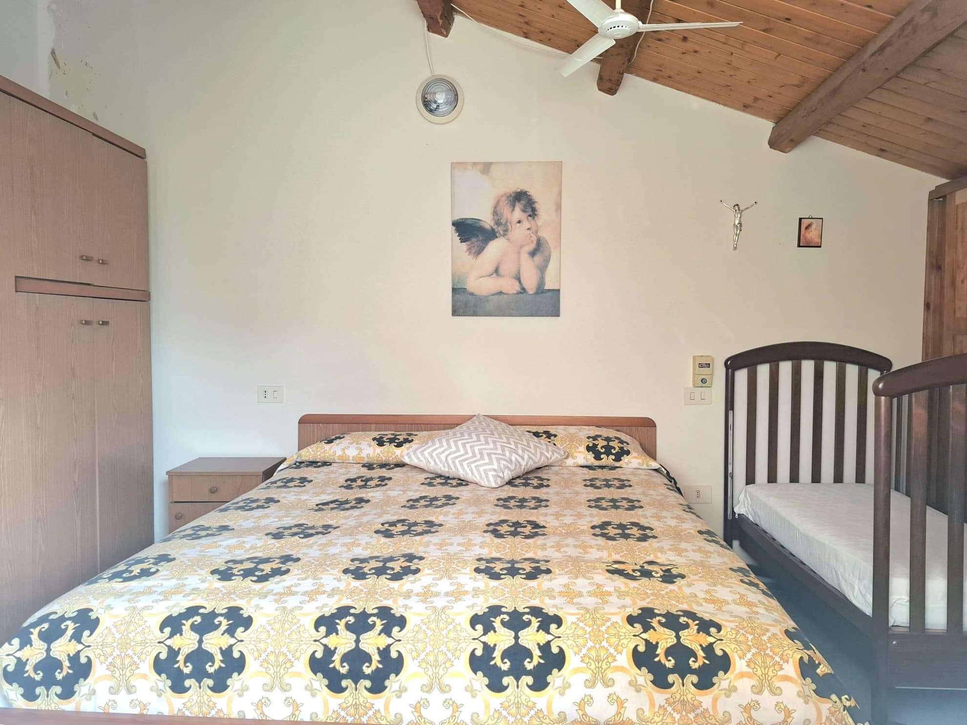 Letto matrimoniale con trapunta a motivi floreali nero e oro, culla per bambino, arredamento in legno e decorazioni religiose, ambiente accogliente in stile rustico.
