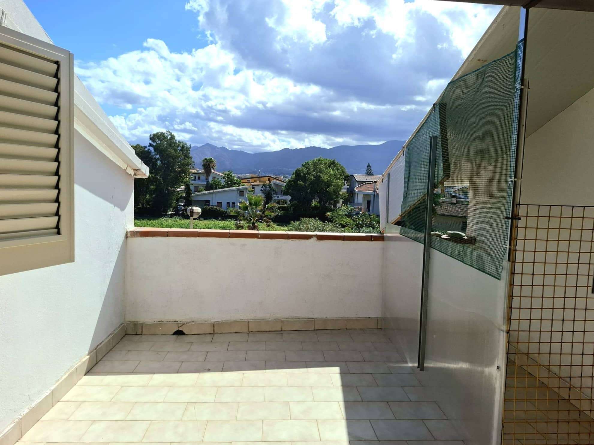 Balcone soleggiato con vista sulle montagne e sul paesaggio urbano, ideale per relax e momenti all'aperto, con pavimento in piastrelle e protezione laterale in rete metallica.