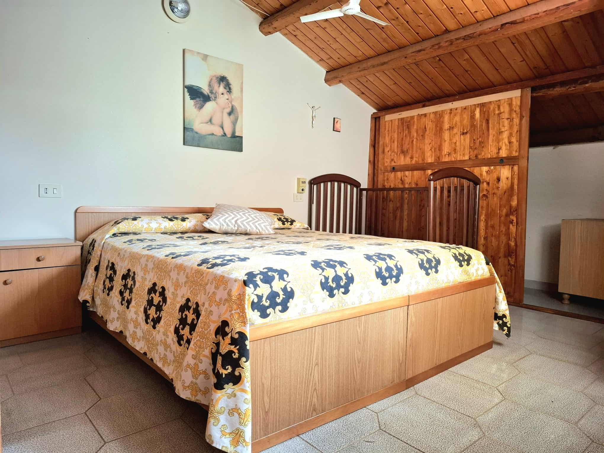 Camera da letto con letto in legno, arredi classici, parete decorata con dipinto di angelo e soffitto in legno, ambiente accogliente e luminoso, ideale per soggiorno tranquillo e rilassante.