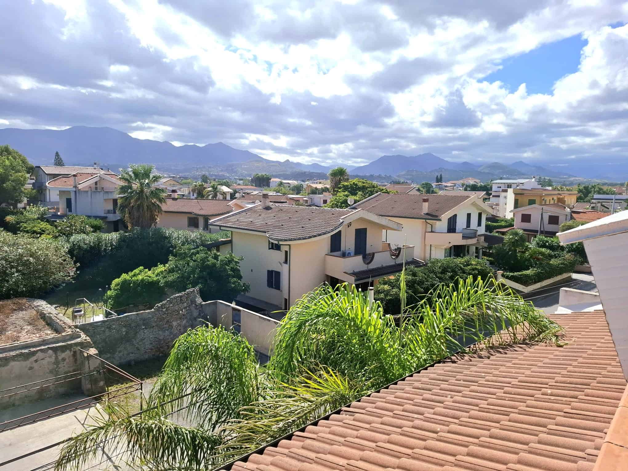 Villetta residenziale con tetto in tegole, vista sul paesaggio urbano e montano, con vegetazione tropicale, case moderne e natura rigogliosa, perfetta per investimento immobiliare a NextCasa.